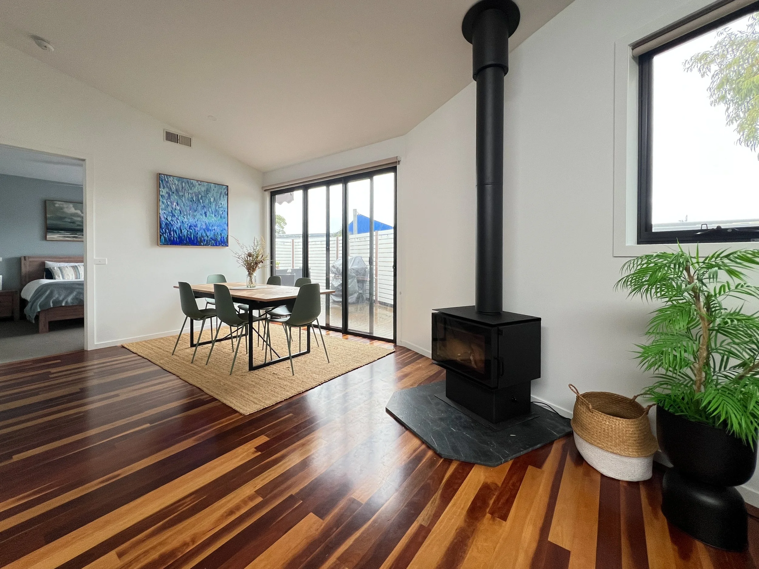 Living room with hardwood floors, a dining area with a table and six chairs, a sliding glass door leading to a balcony, a black stove with a chimney, a potted plant, and artwork on the walls.