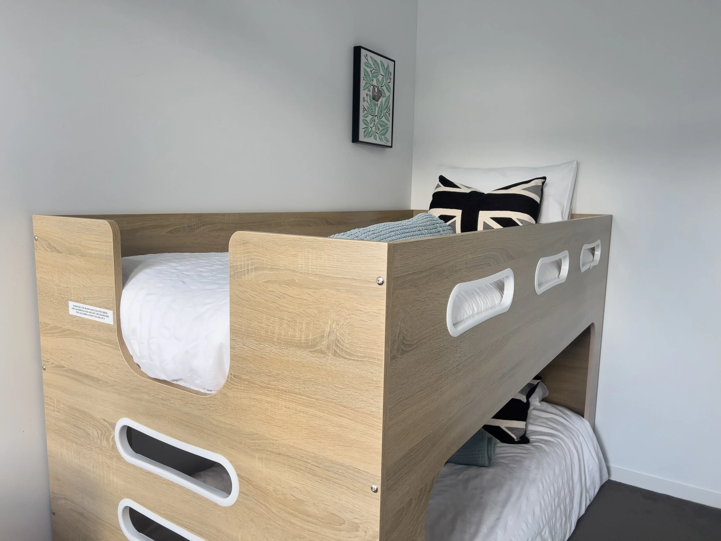 A wooden bunk bed with a light wood finish in a room with white walls. The top bunk has a pillow with a British flag design and a blue blanket, while the bottom bunk has a plain white mirror and a blue pillow. There is a framed artwork of a plant on 