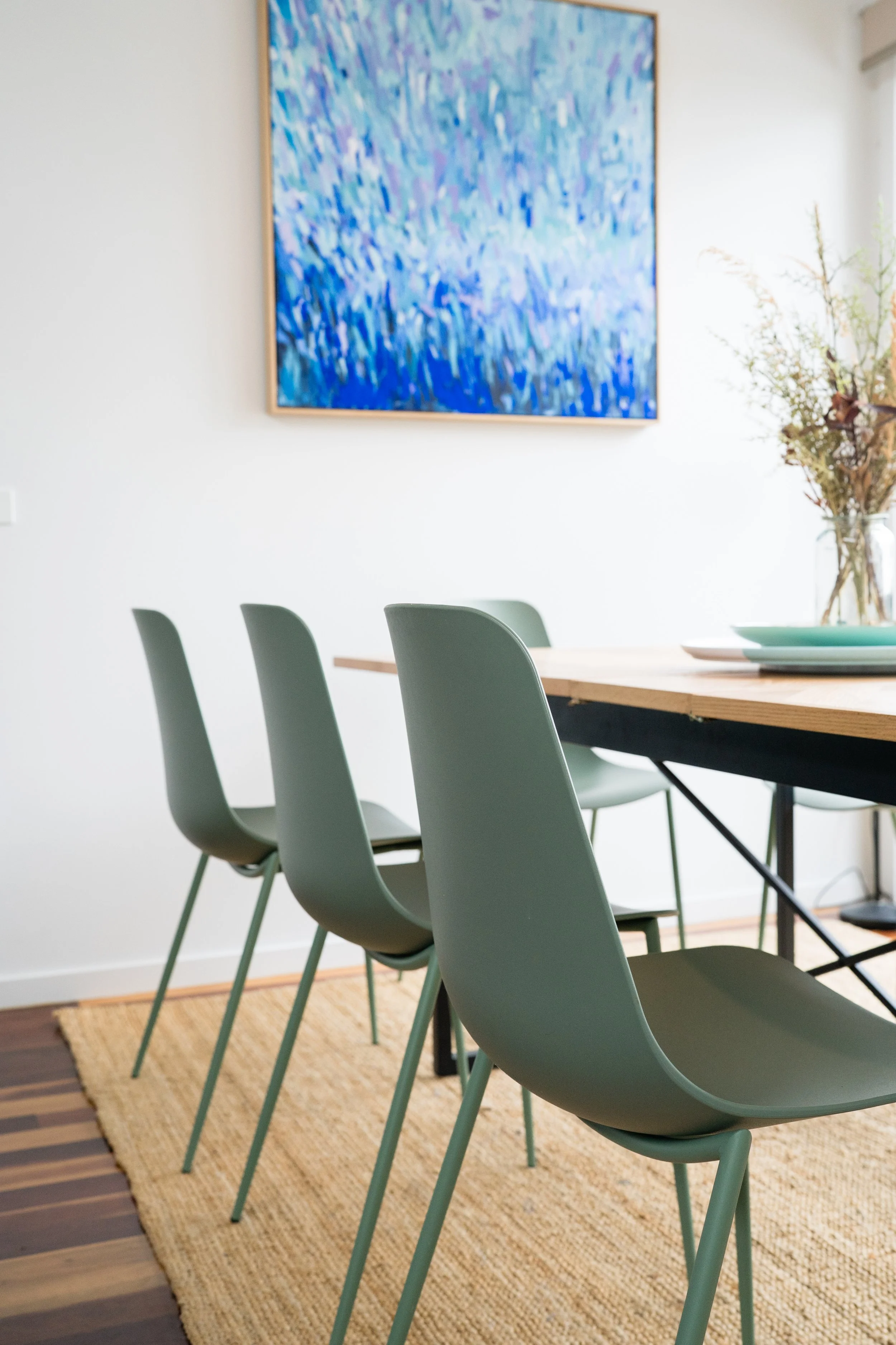 Minimalist dining room with green chairs, a wooden table, a painting with blue abstract art, and a vase with dried plants.