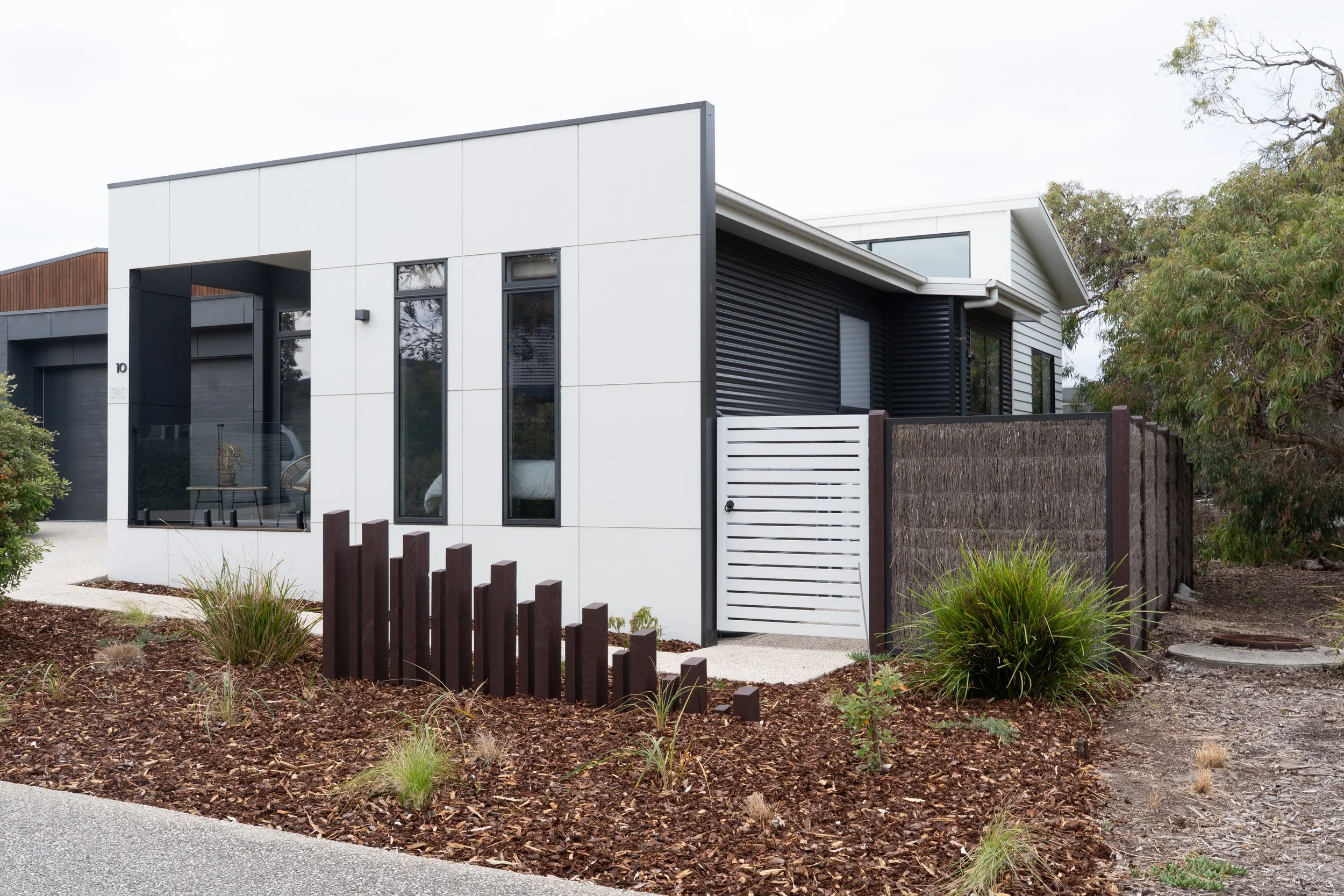 Modern house with white and black exterior, large windows, and surrounding garden with mulch, grass, and plants.