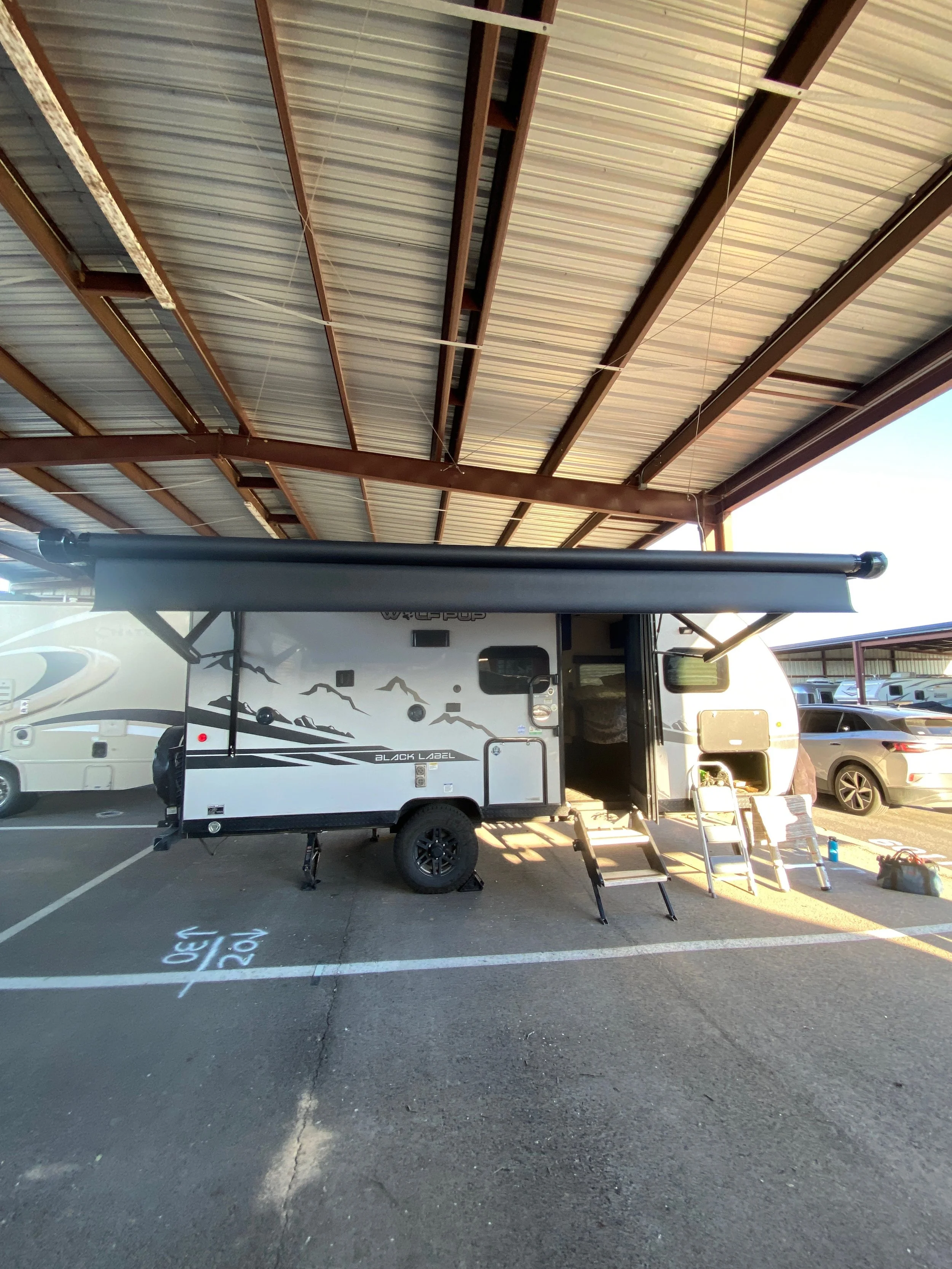 A travel trailer parked in a parking lot under a metal canopy, with an awning extended and steps leading into the door. There are other vehicles parked nearby.