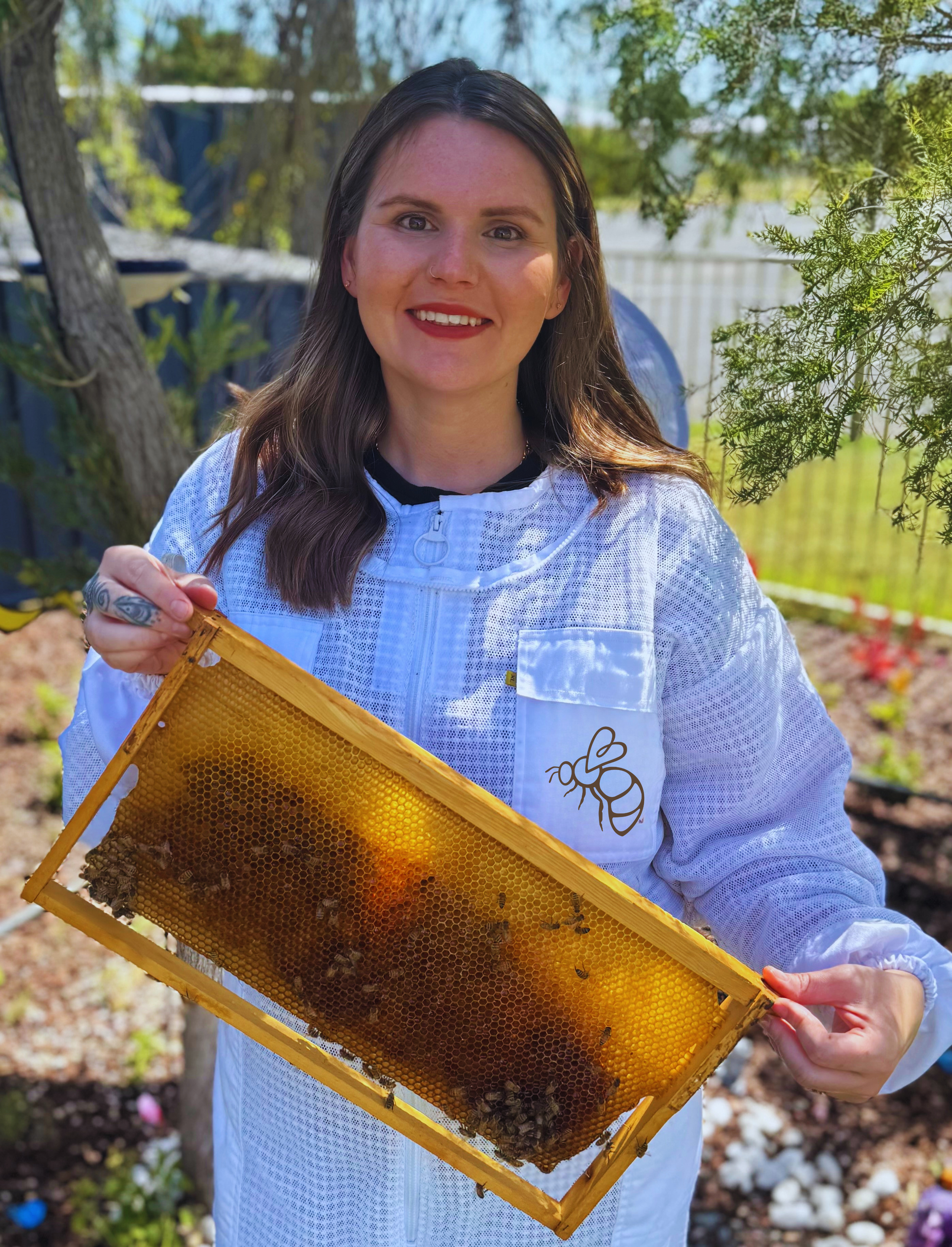 A woman smiling and holding a honey frame in front of beekeeping equipment and wooden bee boxes outdoors in a city setting, with tall buildings in the background.