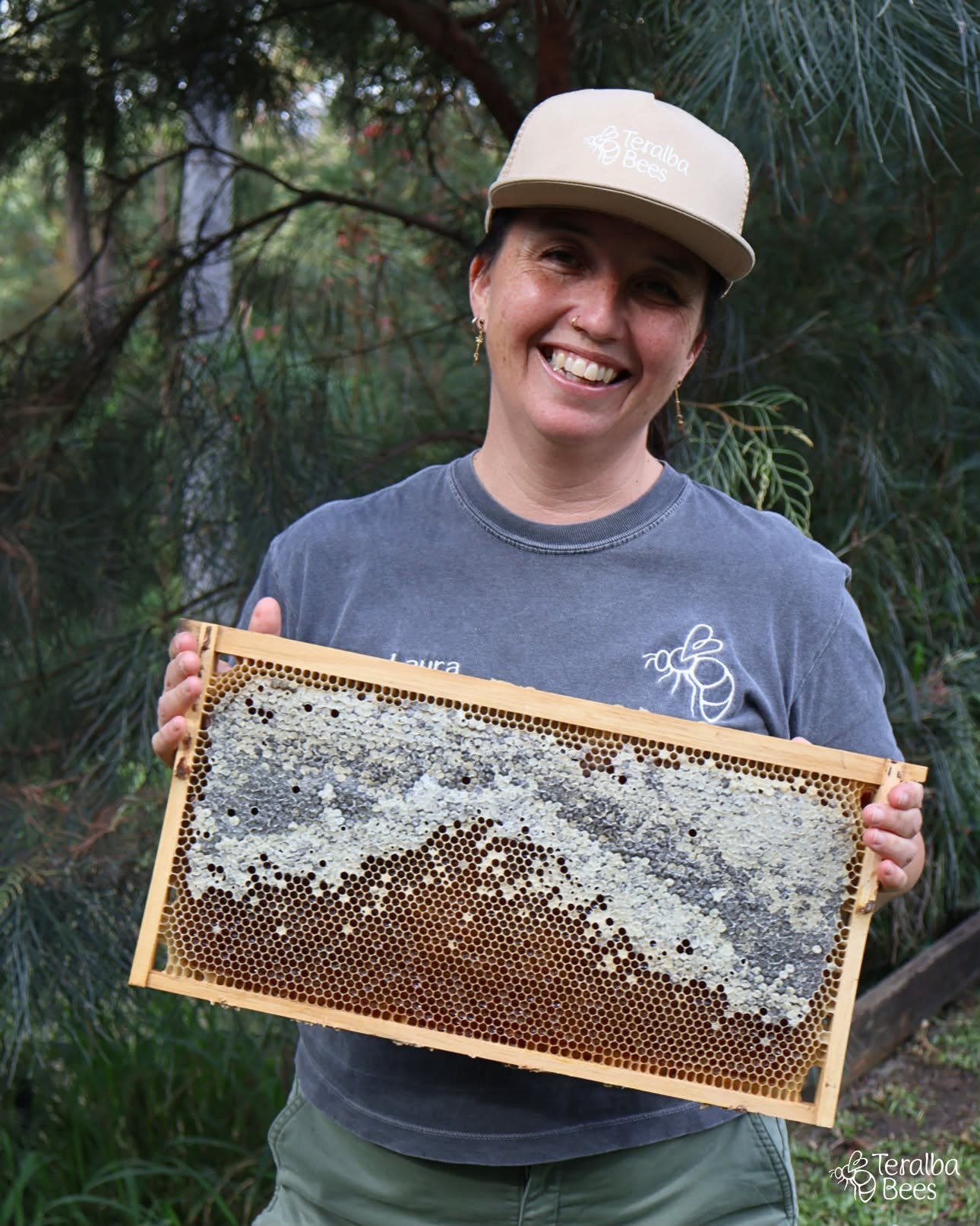 A woman wearing a bee suit standing outdoors next to beehives, smiling at the camera.