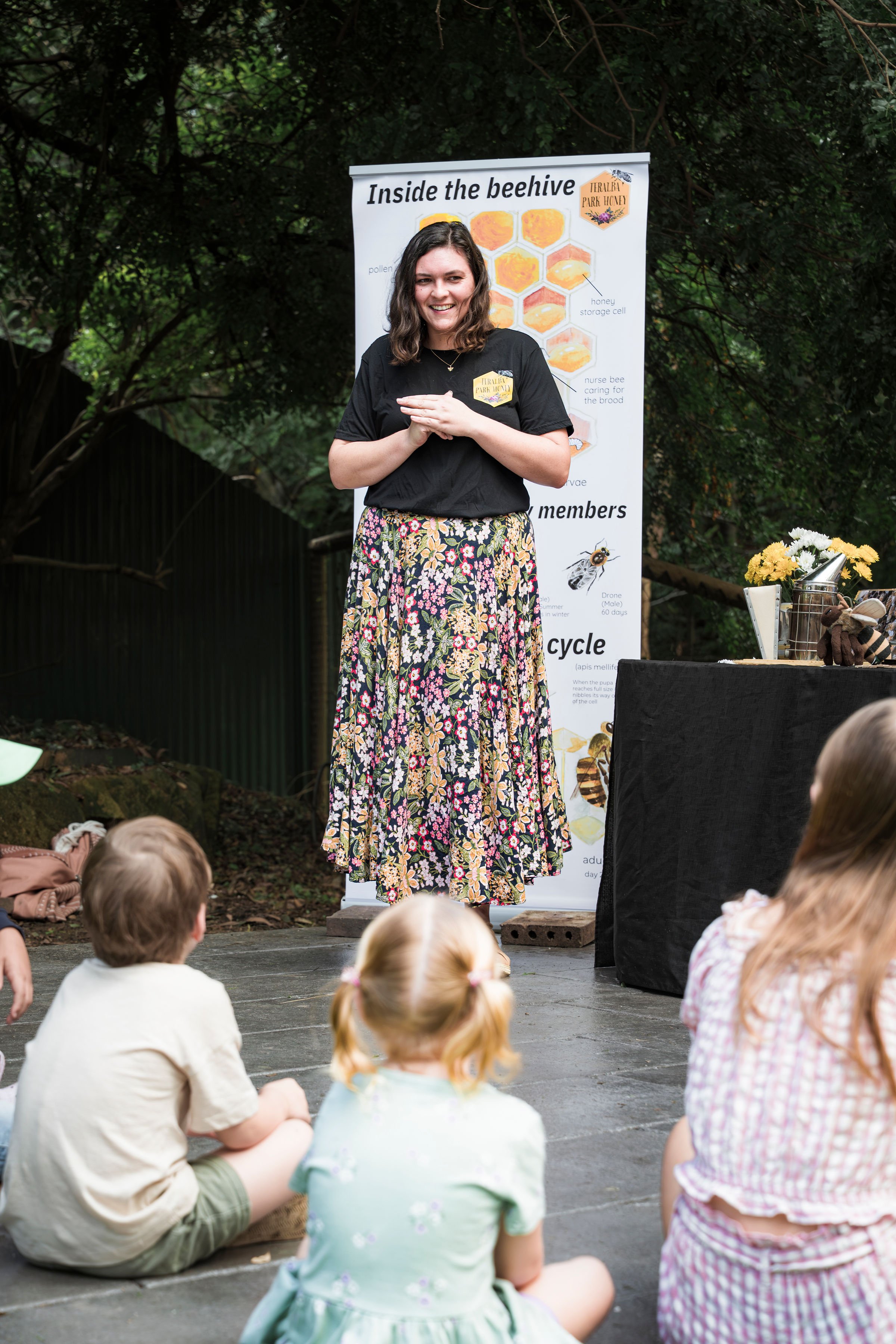 A woman giving a presentation on honey bees outdoors. She stands next to educational posters about honey bee anatomy and hive equipment. A group of children, sitting on the ground, listens attentively.