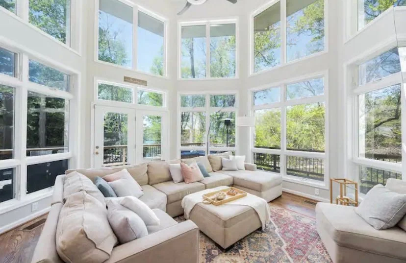 Bright living room with large windows, beige sectional sofa, colorful rug, and outdoor view.