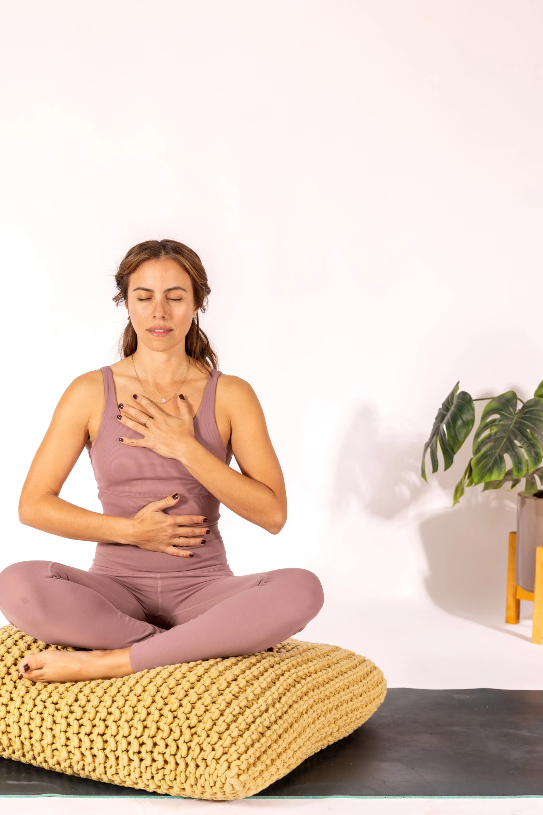 A woman practicing yoga or meditation, sitting cross-legged on a beige cushion in front of a plain white background, with a potted plant on the right side.