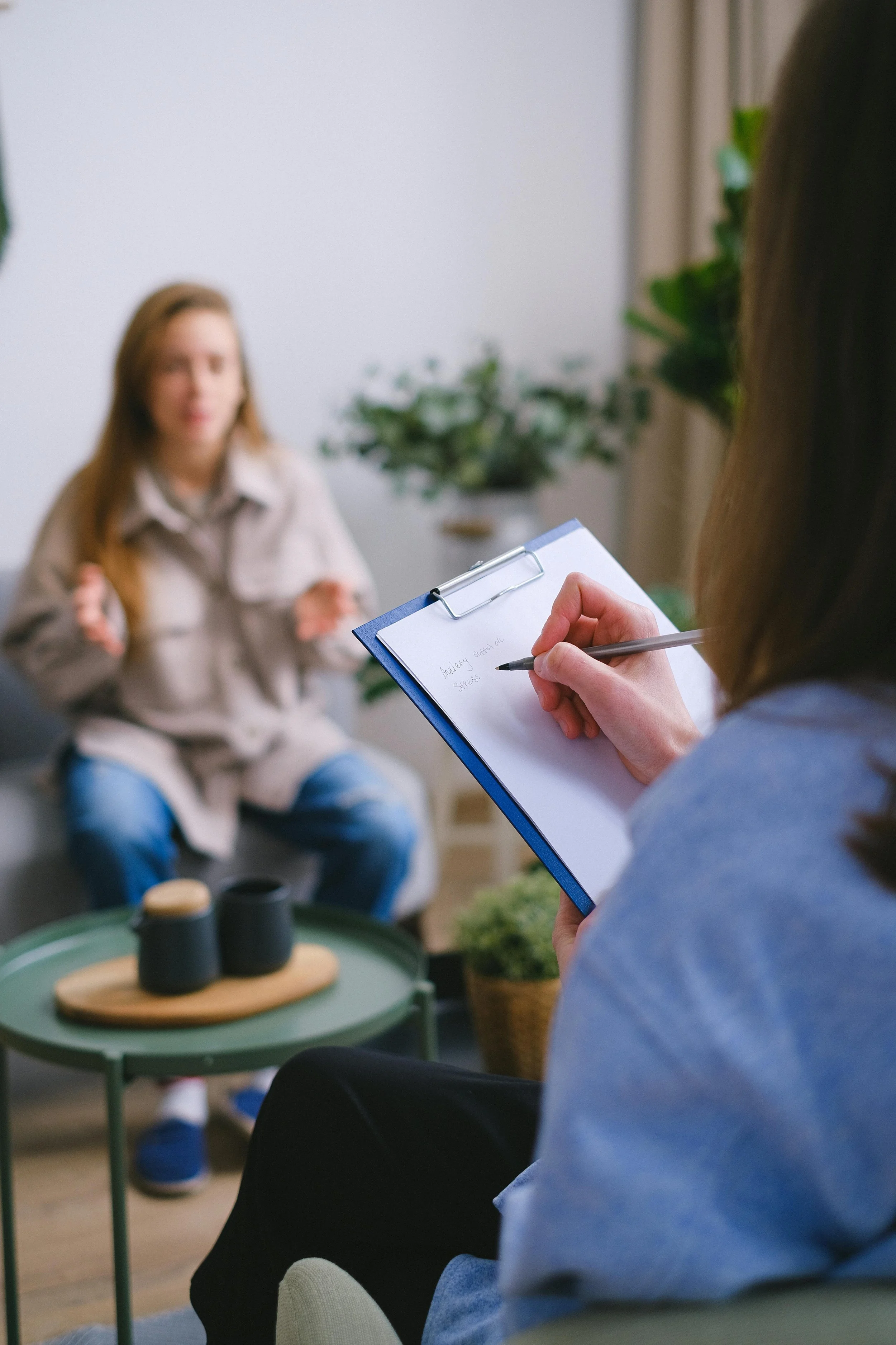 A woman taking notes on a notepad during a therapy session with a young girl in a cozy living room with plants and coffee cups.