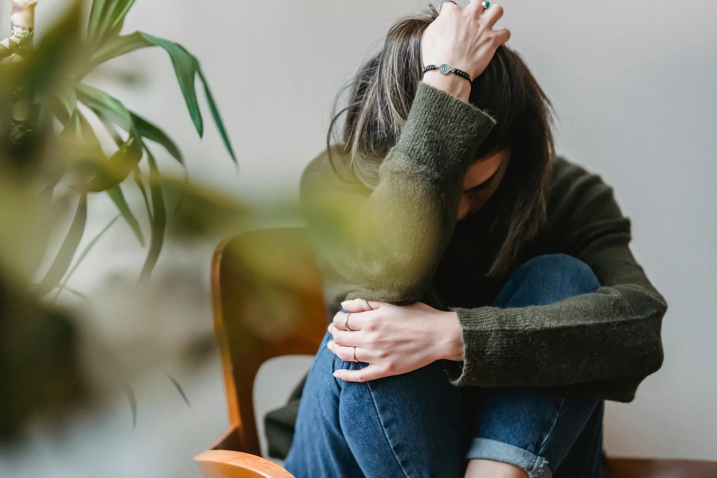 A woman sitting on a wooden chair, holding her head with her left hand and hugging her knees with her right arm, appearing distressed or upset, in a room with a gray wall and some green plants in the foreground.