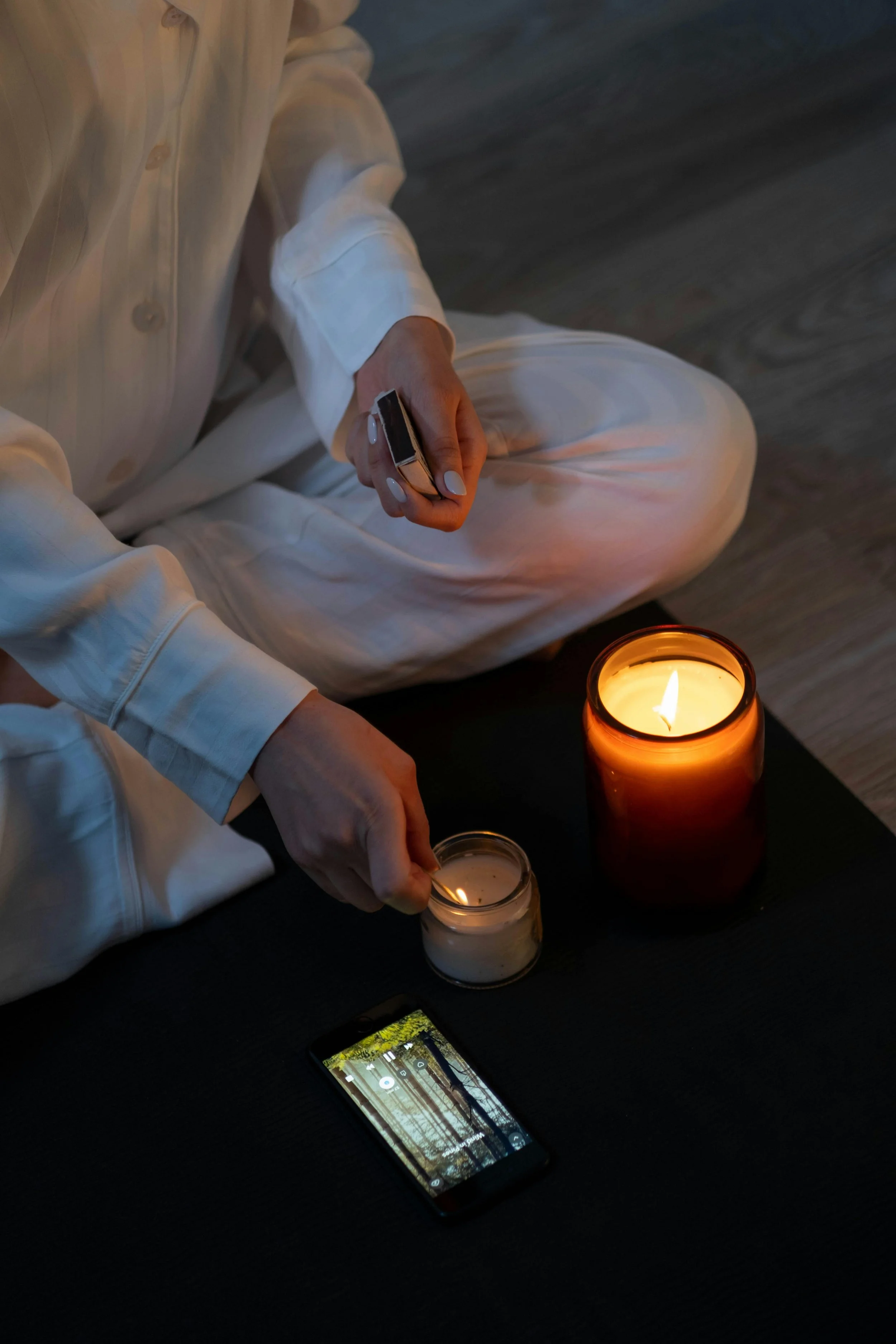 Person dressed in white pajamas sitting on the floor, lighting candles with a matchstick, with a smartphone nearby on a dark surface, and a large lit candle in a brown jar.