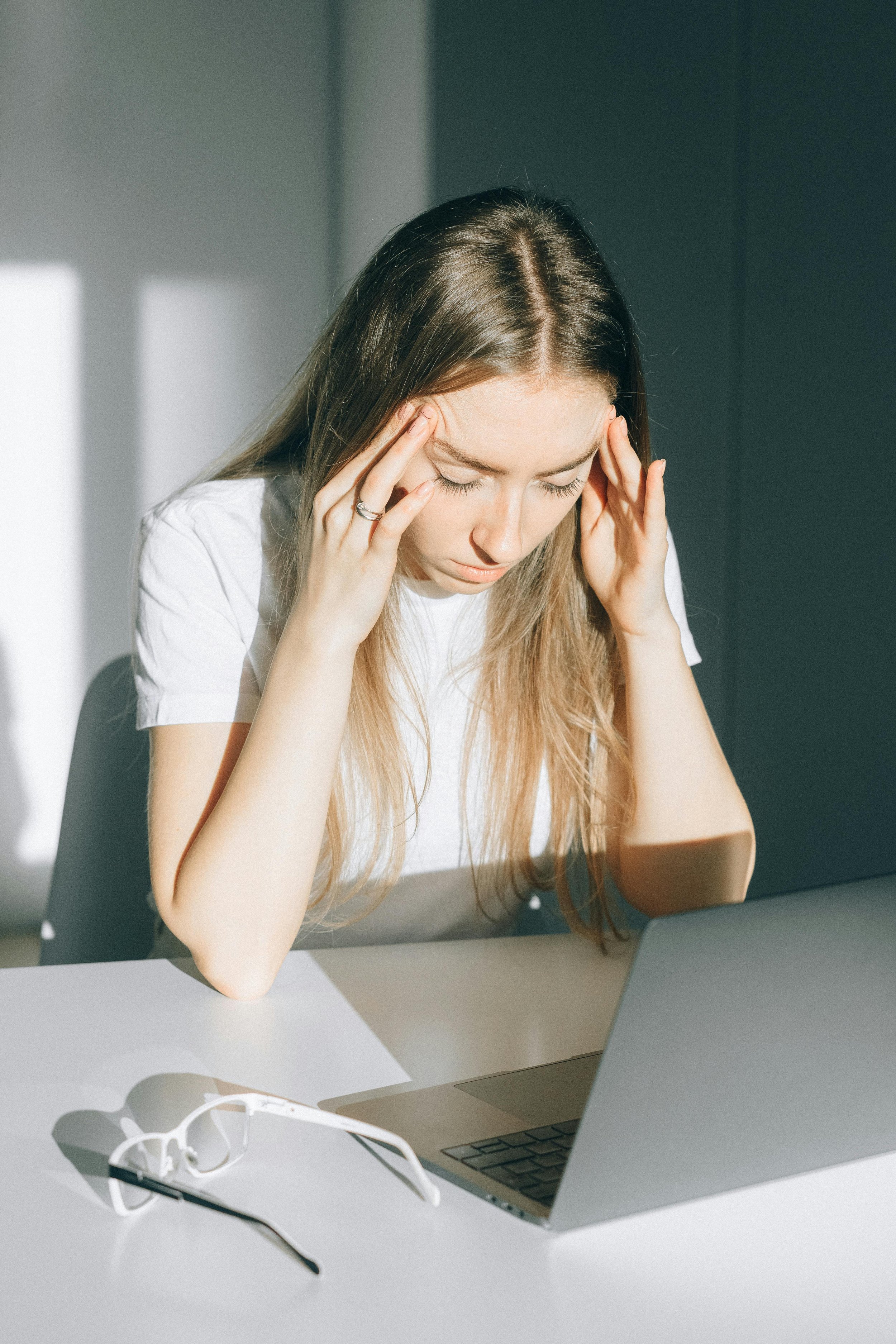 Young woman sitting at a desk, holding her head in stress, with a laptop and eyeglasses on the table.
