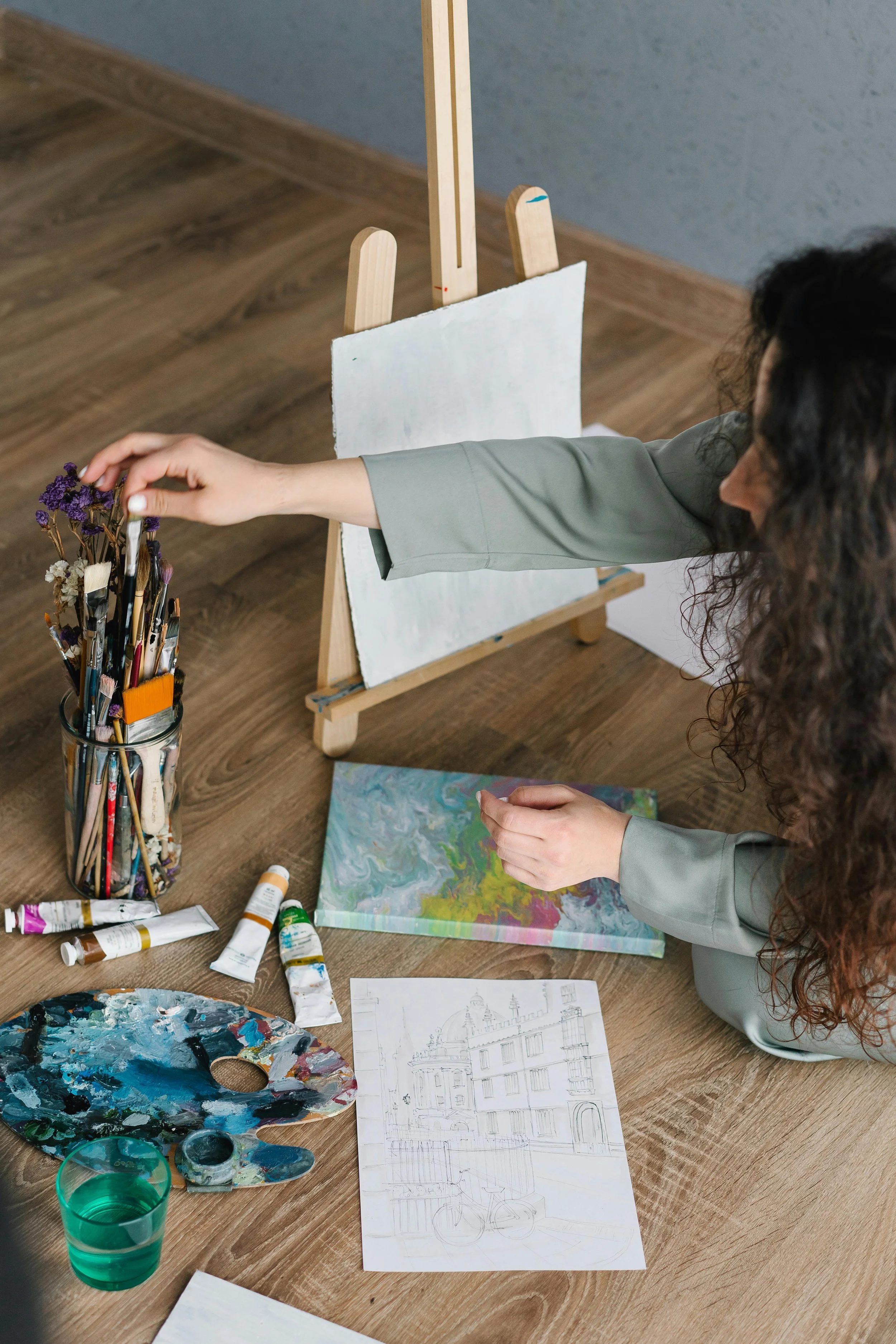 A woman is arranging paintbrushes in a jar on a wooden table with art supplies, including paint tubes, a paint palette, a glass of water, and sketches of buildings and a bicycle. An easel with a blank canvas is in the background.