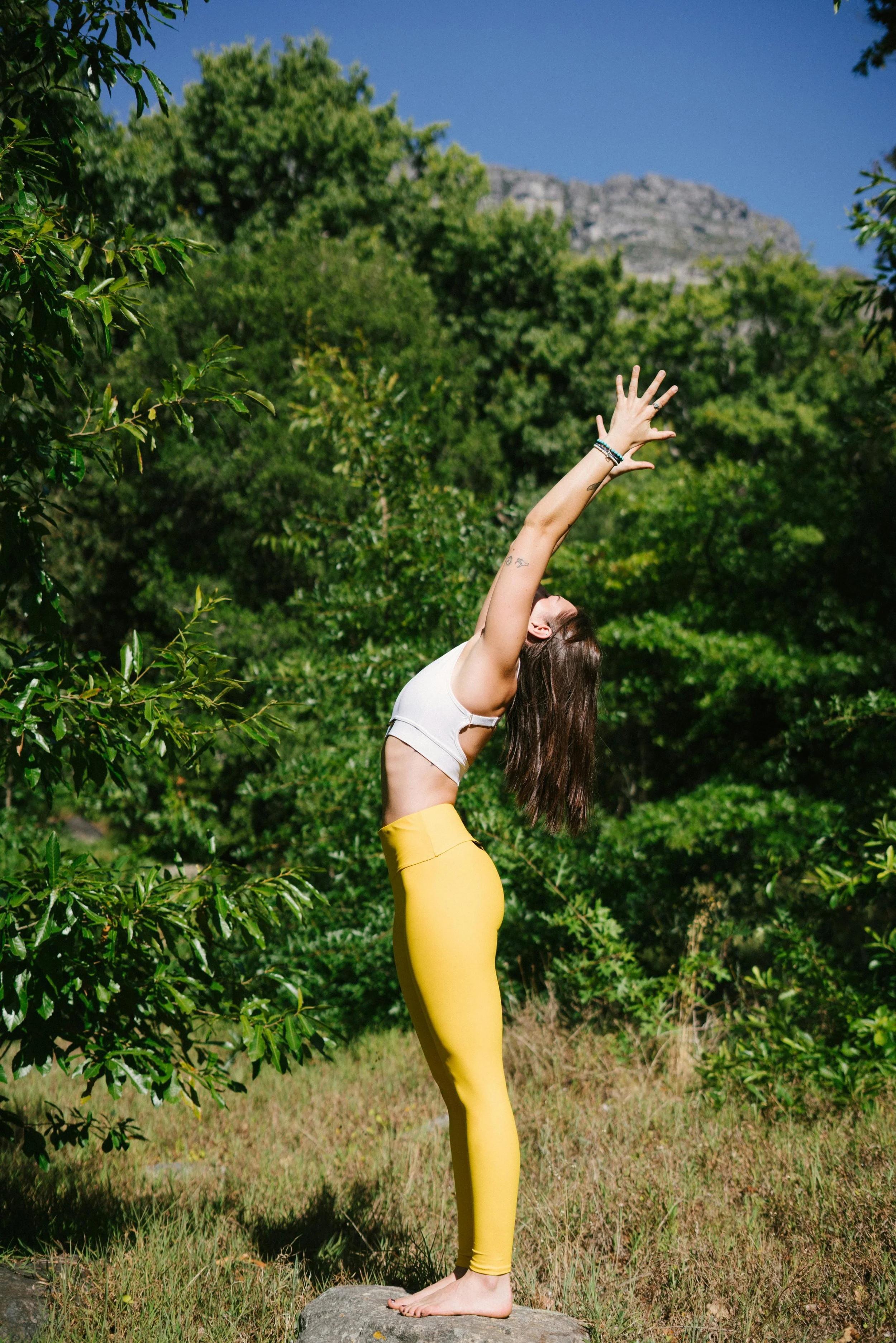 A woman practicing yoga outdoors on a sunny day, standing on a rock with arms extended overhead, surrounded by green trees and a mountain in the background.
