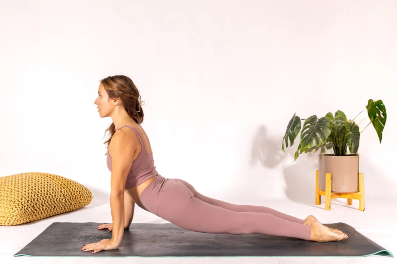 A woman practicing yoga in a yoga pose called the upward dog on a black mat in a room with minimalist decor, a yellow knit pillow, and a potted Monstera plant.