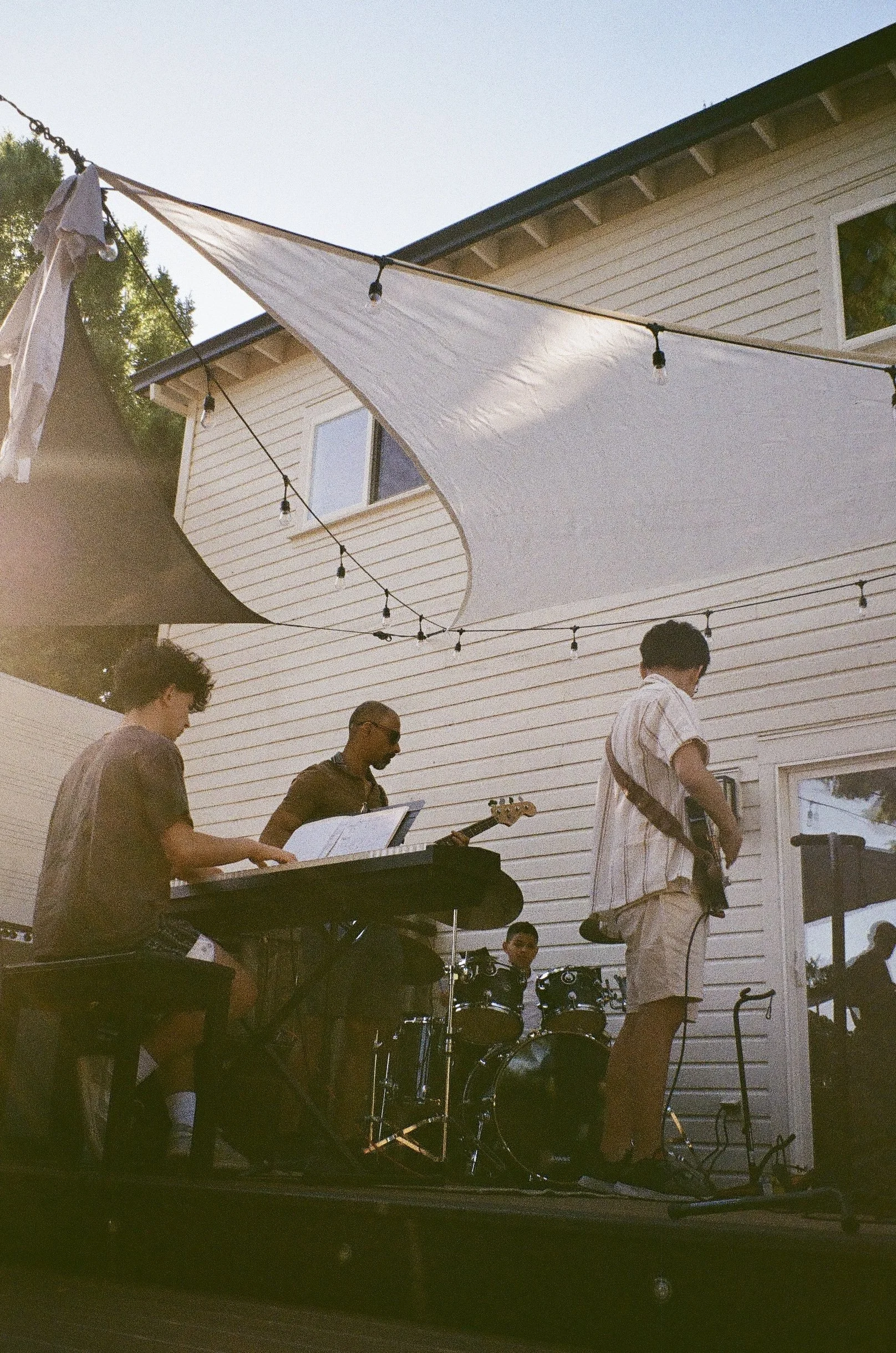 A group of musicians playing instruments outdoors in a backyard, with a white house and string lights overhead, during daytime.