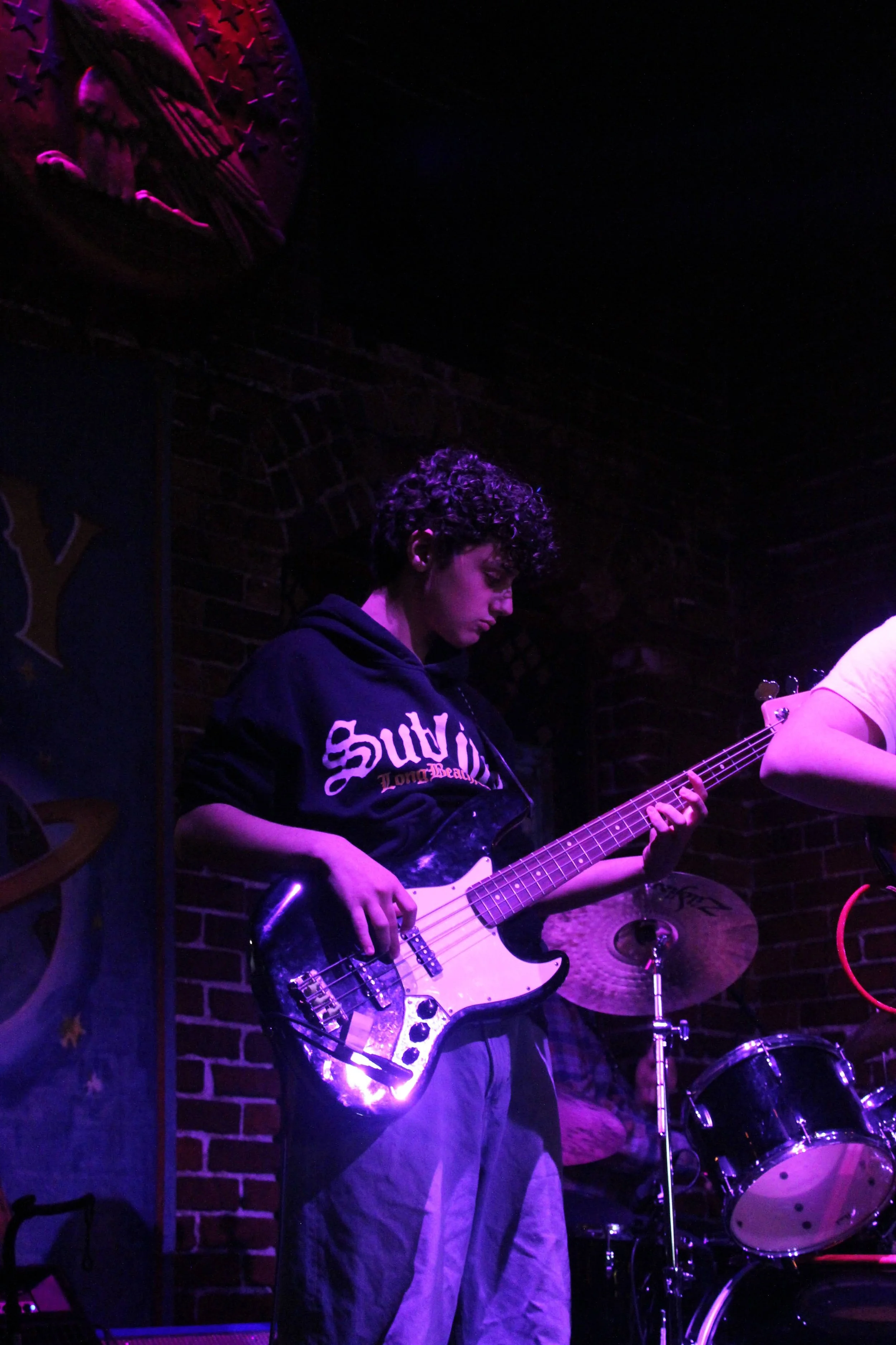 A young man with curly hair playing an electric bass guitar on stage, illuminated by purple stage lights, with a brick wall background and a drum set nearby.