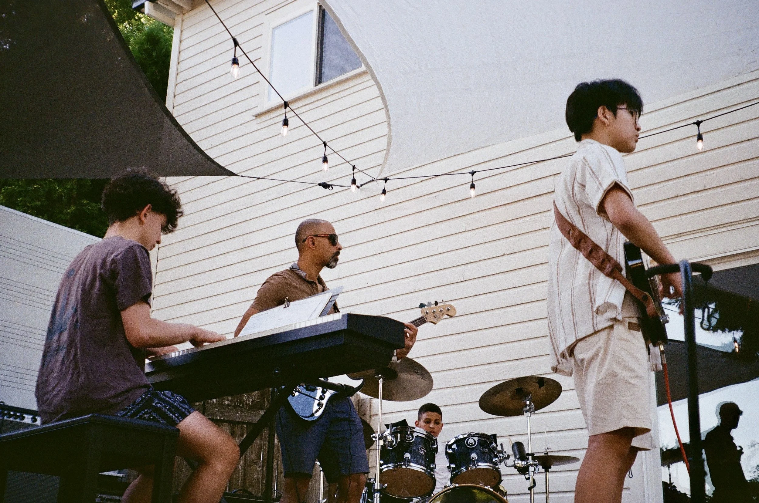 A group of children and an adult playing musical instruments outdoors in front of a white building, with string lights overhead.