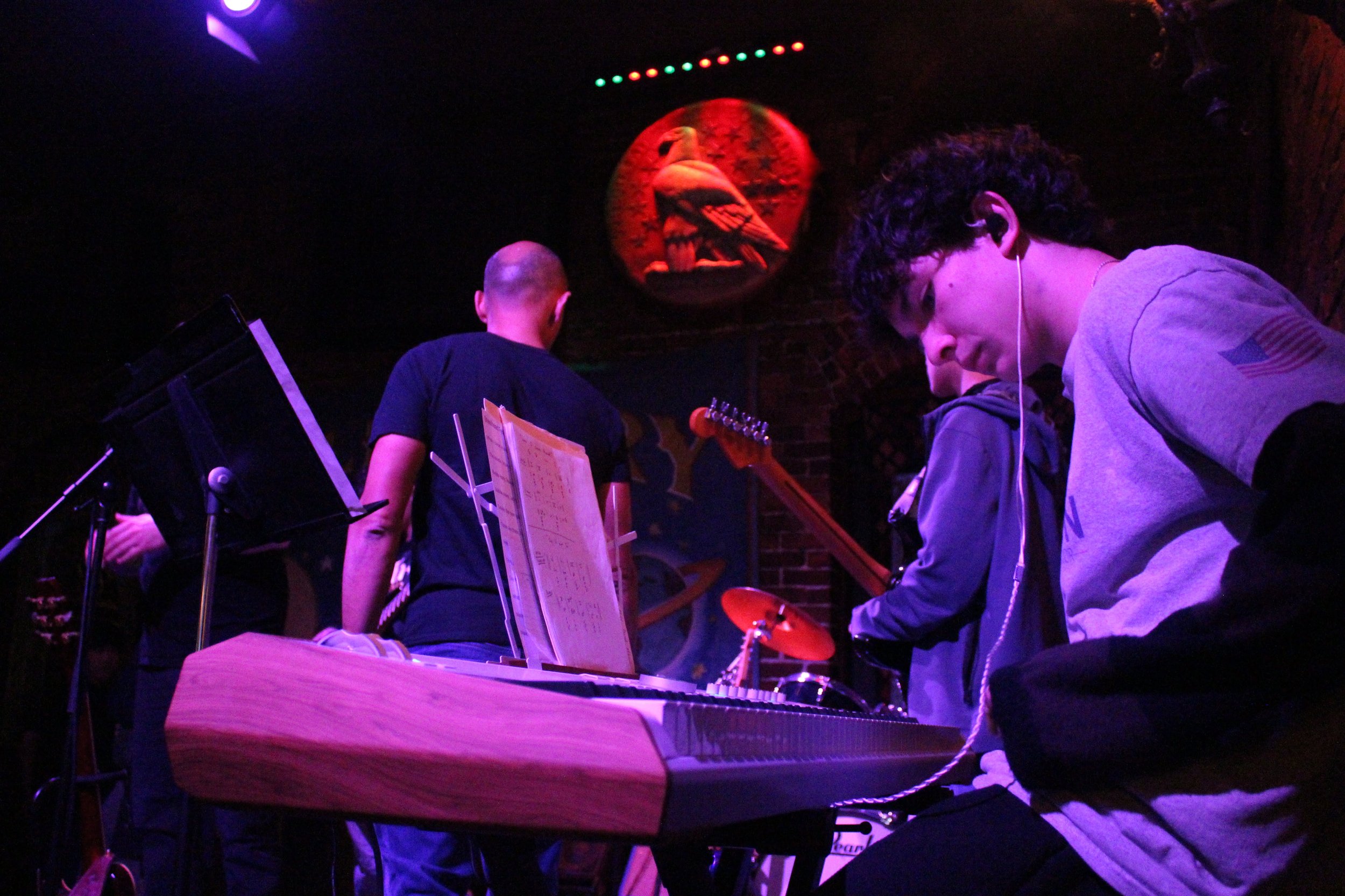A jazz band performing live on stage in a dimly lit venue with purple and red lighting, featuring a pianist, a guitarist, and a saxophonist, with a spotlight on the saxophonist.