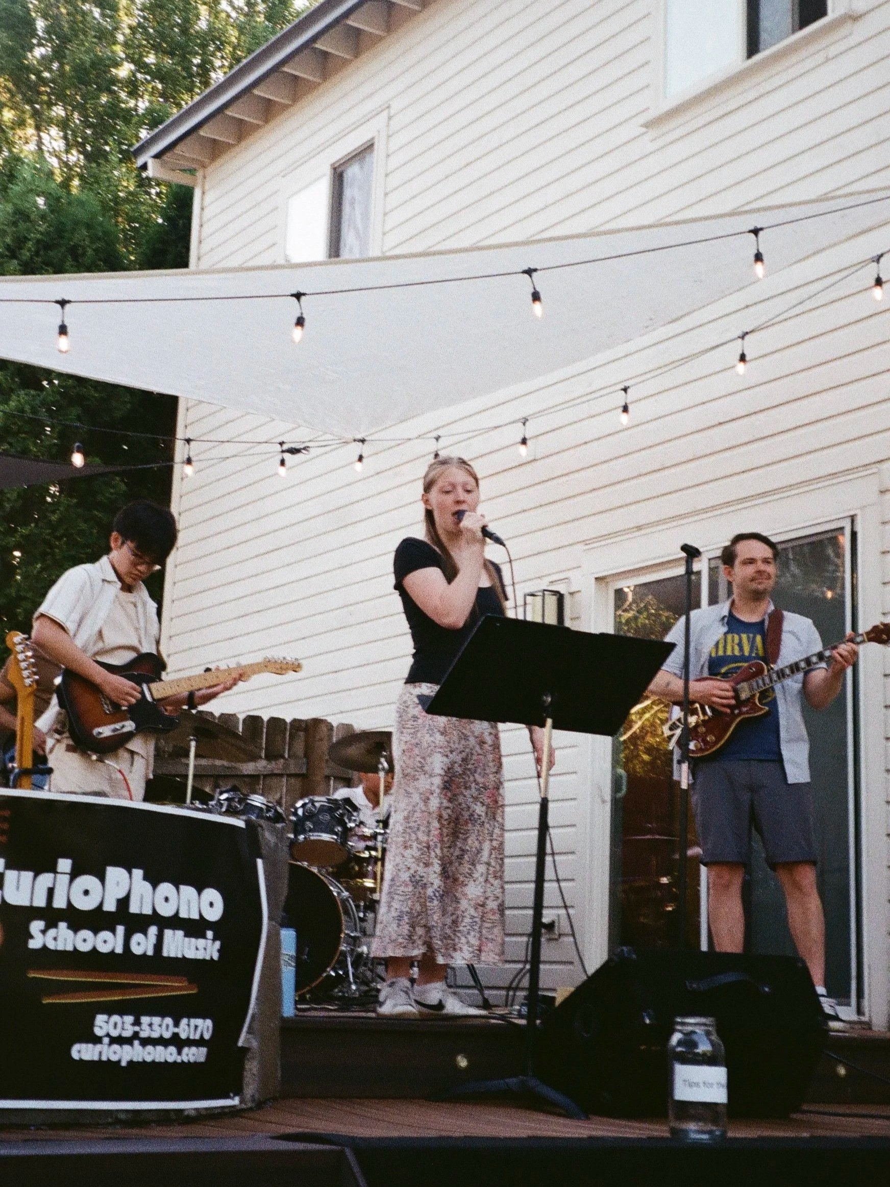A live outdoor musical performance by a group of four young musicians, including a female singer with a microphone, a guitarist, and a bassist, on a wooden deck with string lights overhead and a white house with windows in the background. There is a sign in the foreground that reads 'School of Music' with contact information.