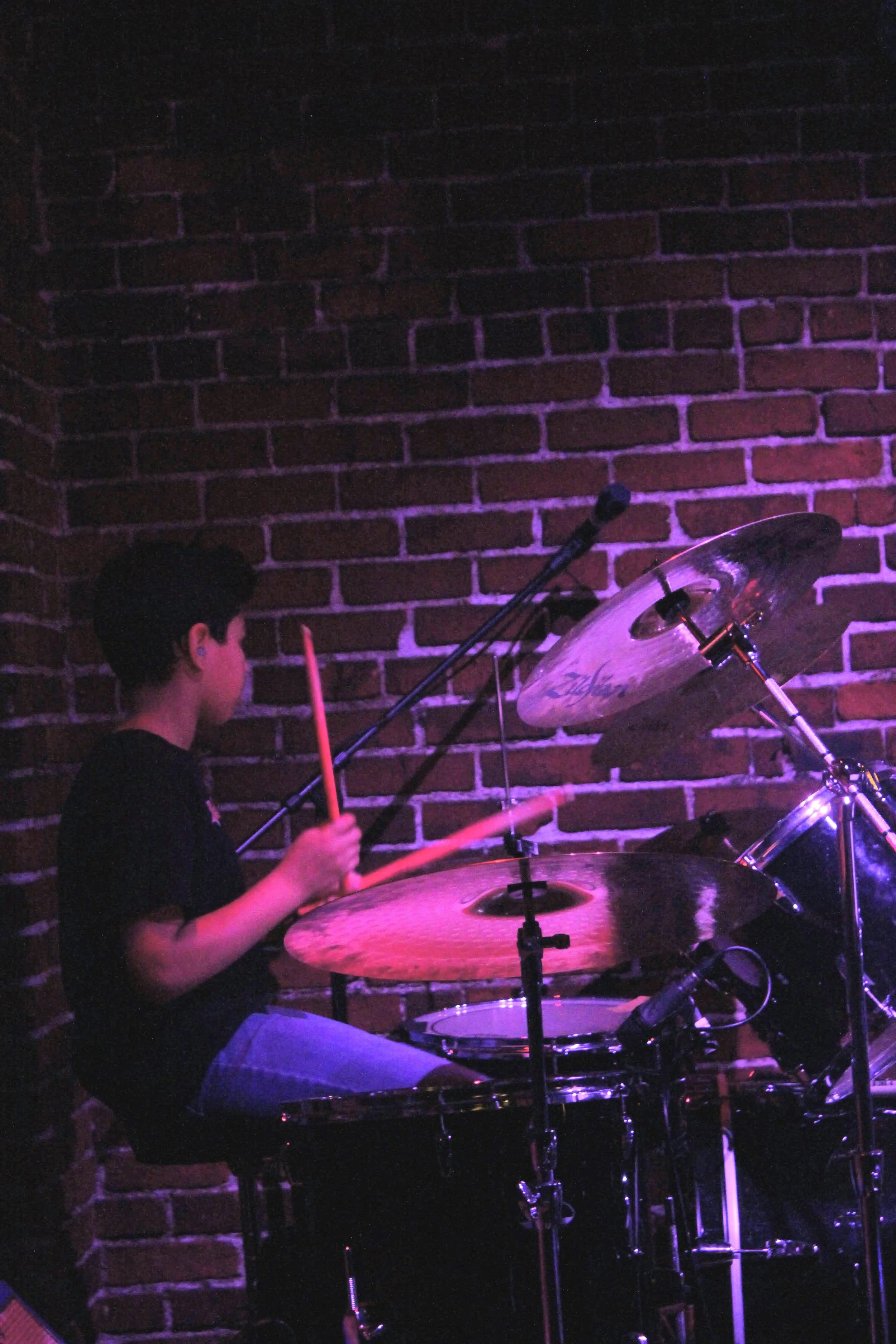 A young boy playing drums on stage against a brick wall, with purple stage lighting.