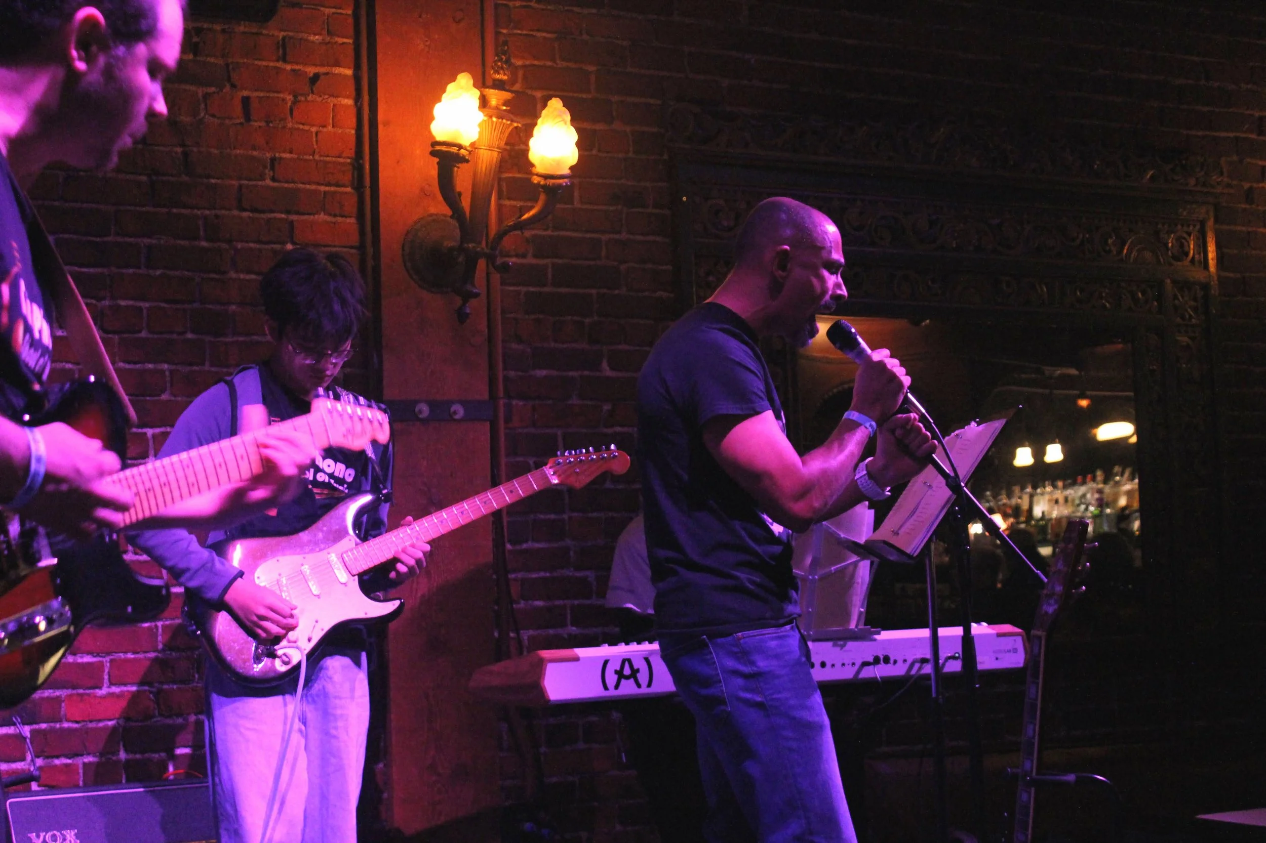 A group of musicians performing on stage in a dimly lit venue with brick walls and warm lighting, including a guitarist and a singer.
