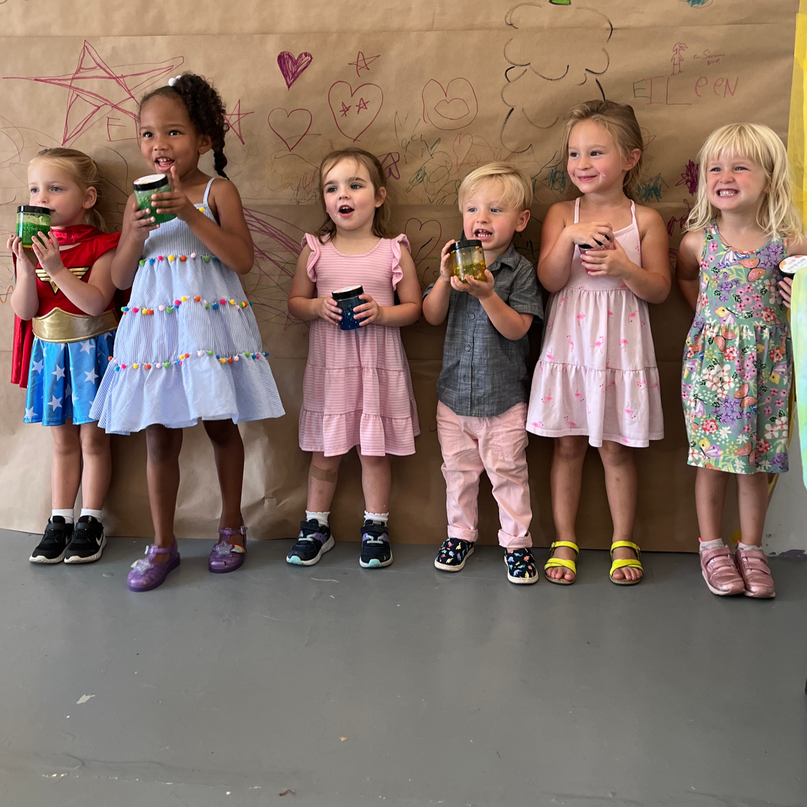 Children dressed in colorful costumes standing in front of a large paper backdrop with hand-drawn illustrations and the words "DRAW ON ME" on it.