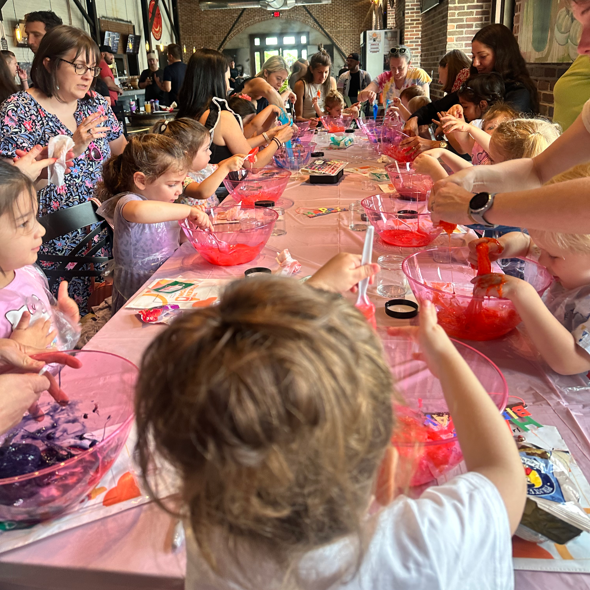 Children and adults gathered around a long table with pink tablecloths, engaging in arts and crafts activities, with some items and supplies on the table, in an indoor setting with exposed brick walls and industrial ceiling.