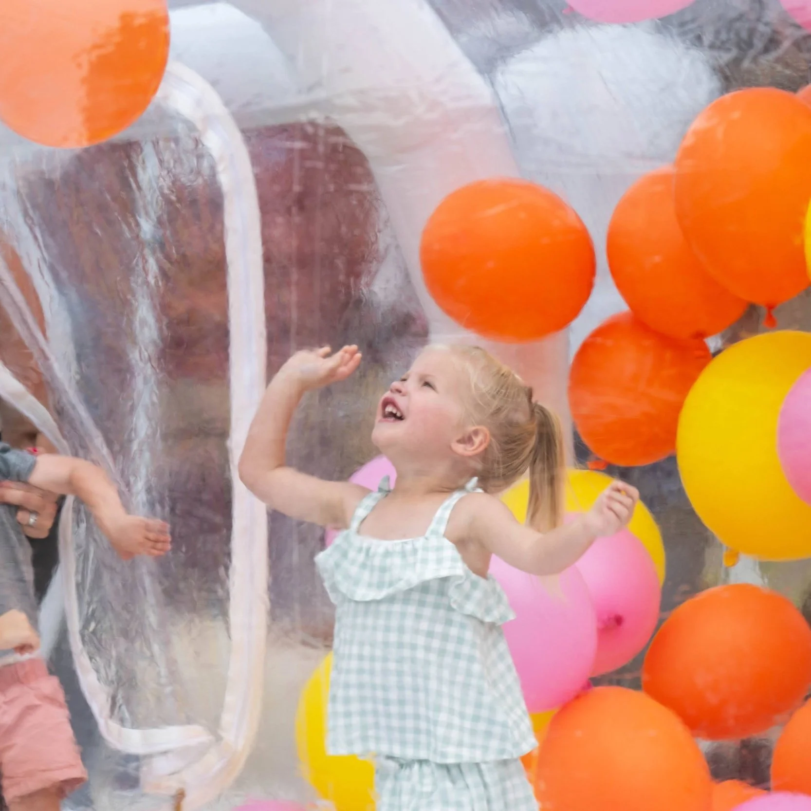 A young girl in a sleeveless gingham dress playing surrounded by colorful balloons and a clear plastic tunnel.