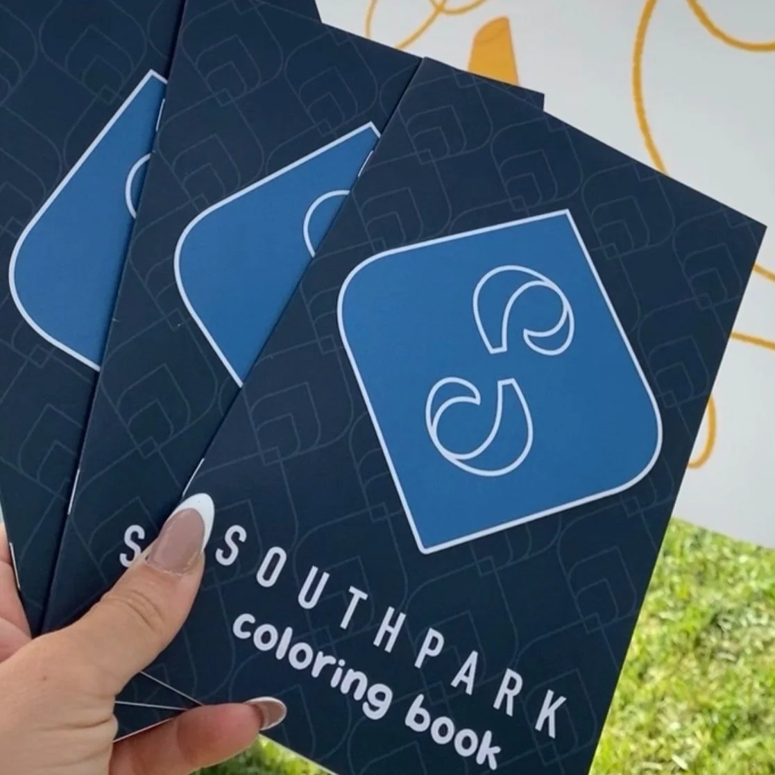 Four South Park-themed coloring books with blue covers being held by a person, with a background showing grass and a white wall with yellow lines.