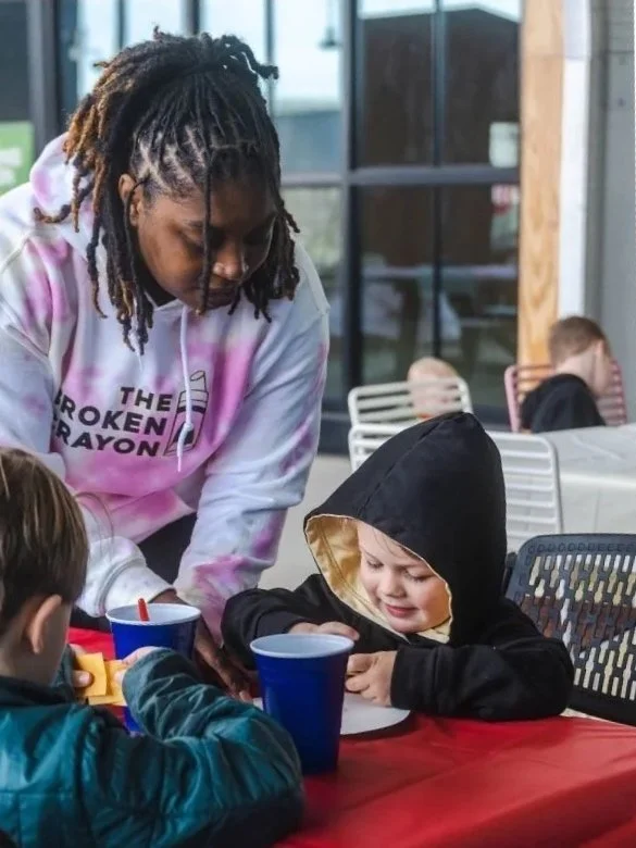 An adult woman with dreadlocks and a pastel tie-dye hoodie leaning over a table to speak with two children sitting at a table with red tablecloths. The children are eating and drinking from blue cups, with one child wearing a hooded jacket and the other a teal jacket, and they are engaged in a conversation or activity.