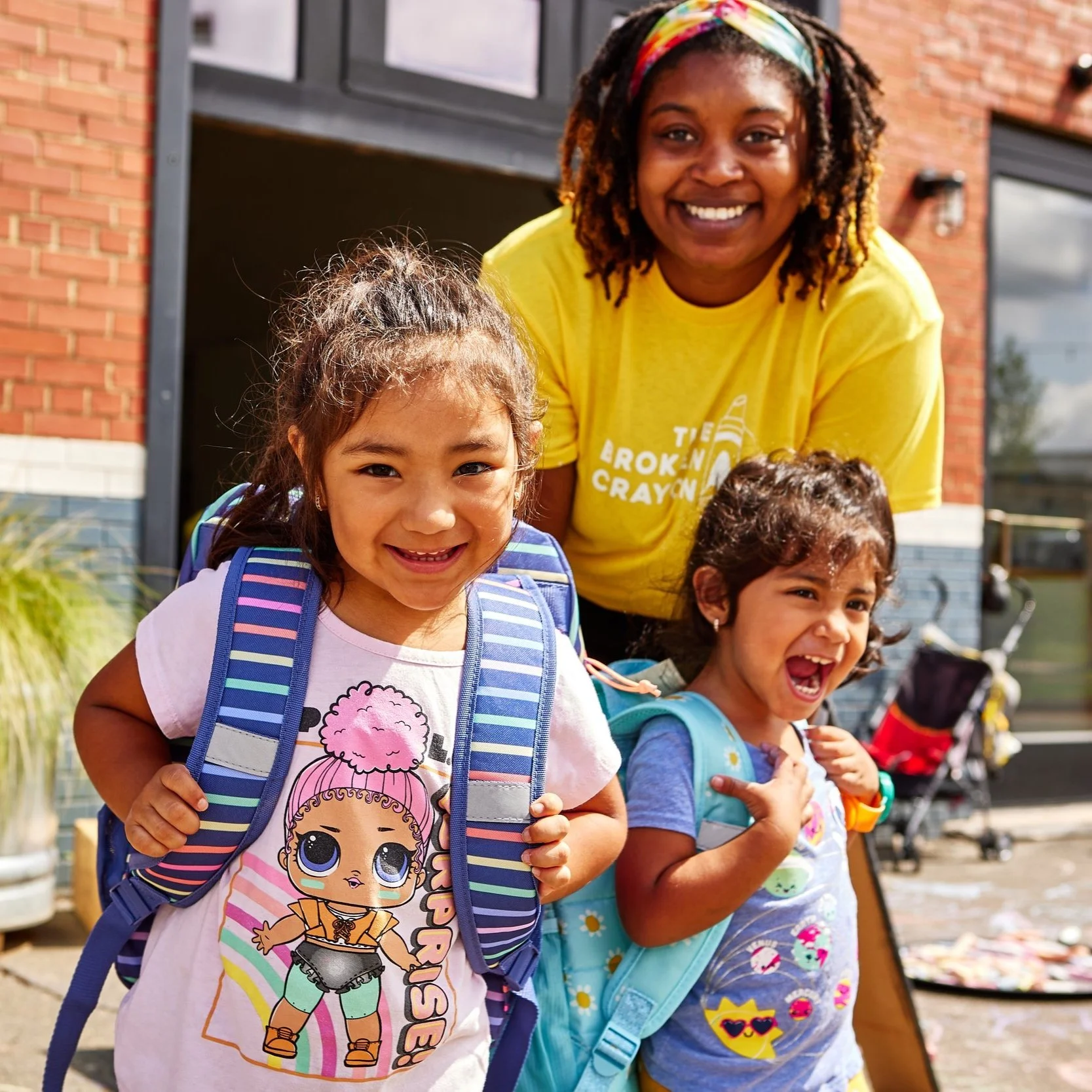 A woman and two young girls smiling outdoors in front of a brick building. The woman is wearing a yellow T-shirt and a colorful headband, while the girls have backpacks and are showing expressions of happiness.