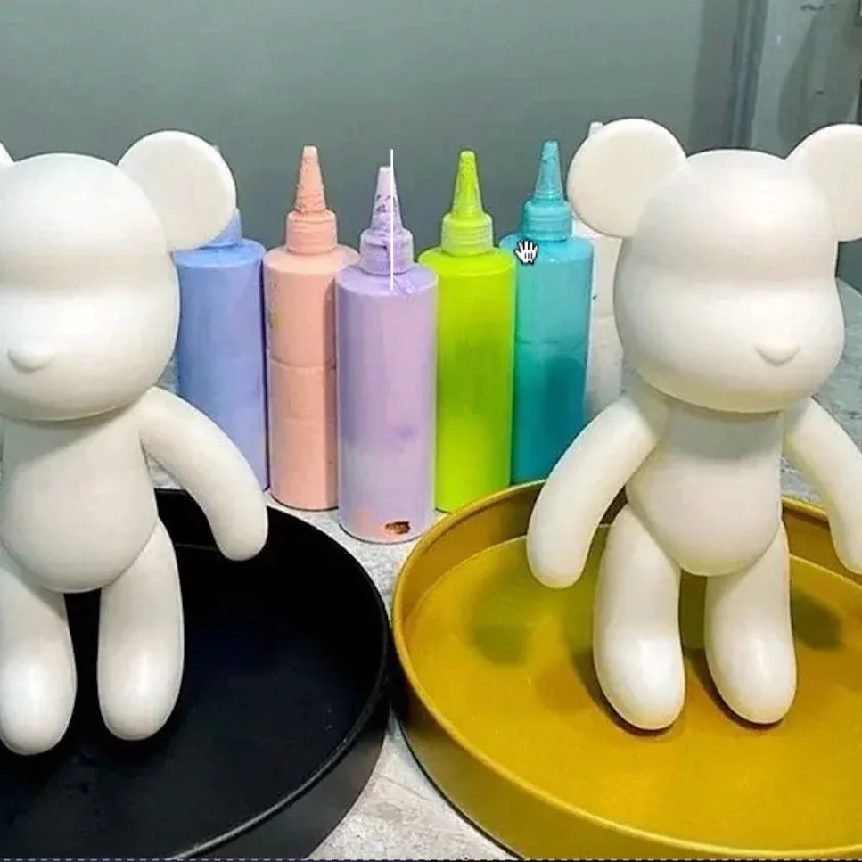 Two white bear-shaped figurines with round ears, one on a black tray and one on a yellow tray, with colorful bottles of paint or glue in the background.