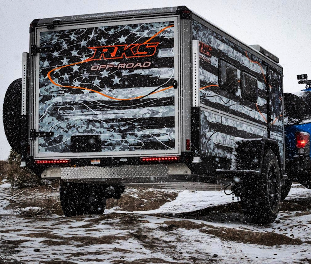 Off-road camper trailer with the RKG Off-Road logo, parked on snowy terrain during snowfall.