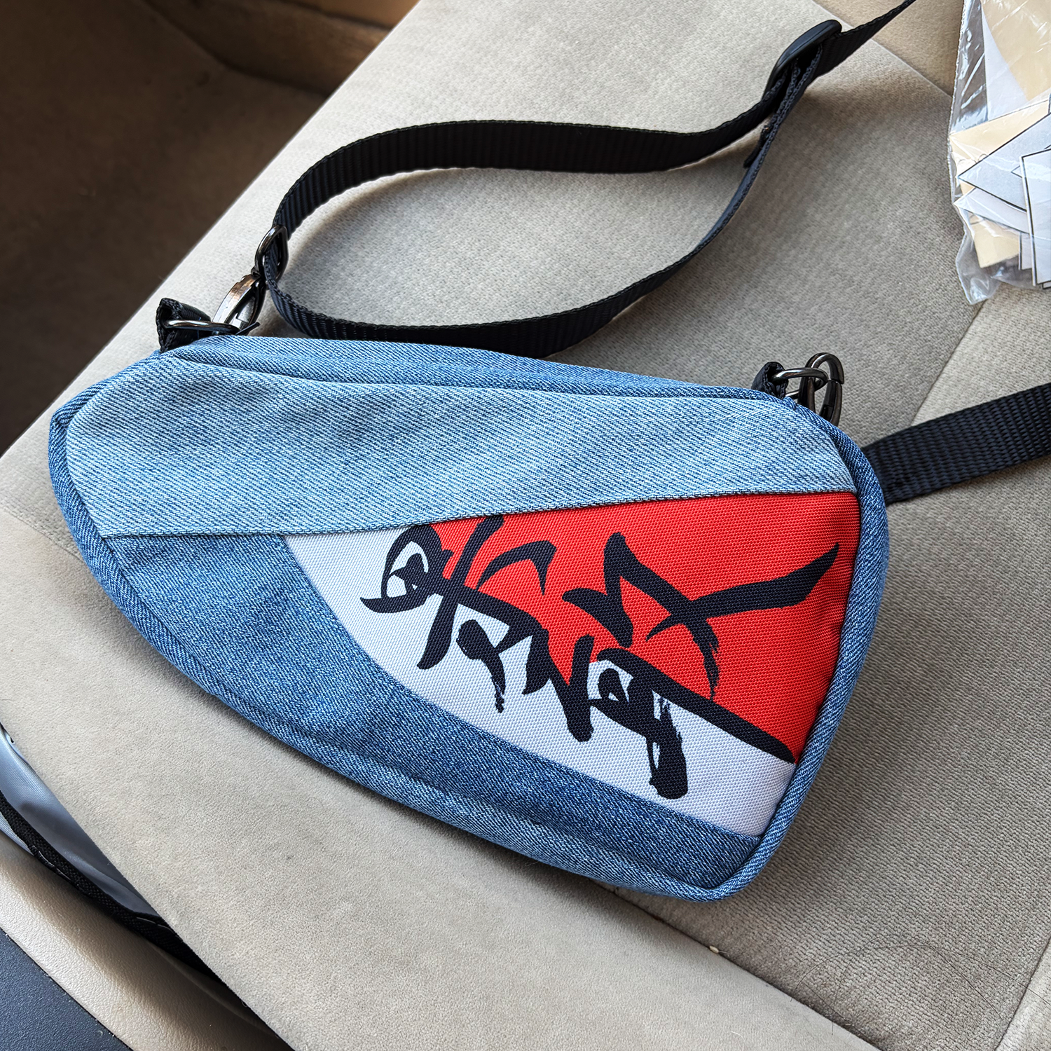 A denim style crossbody bag with a graffiti-style graphic, lying on a beige upholstered surface. The bag has a black adjustable strap.