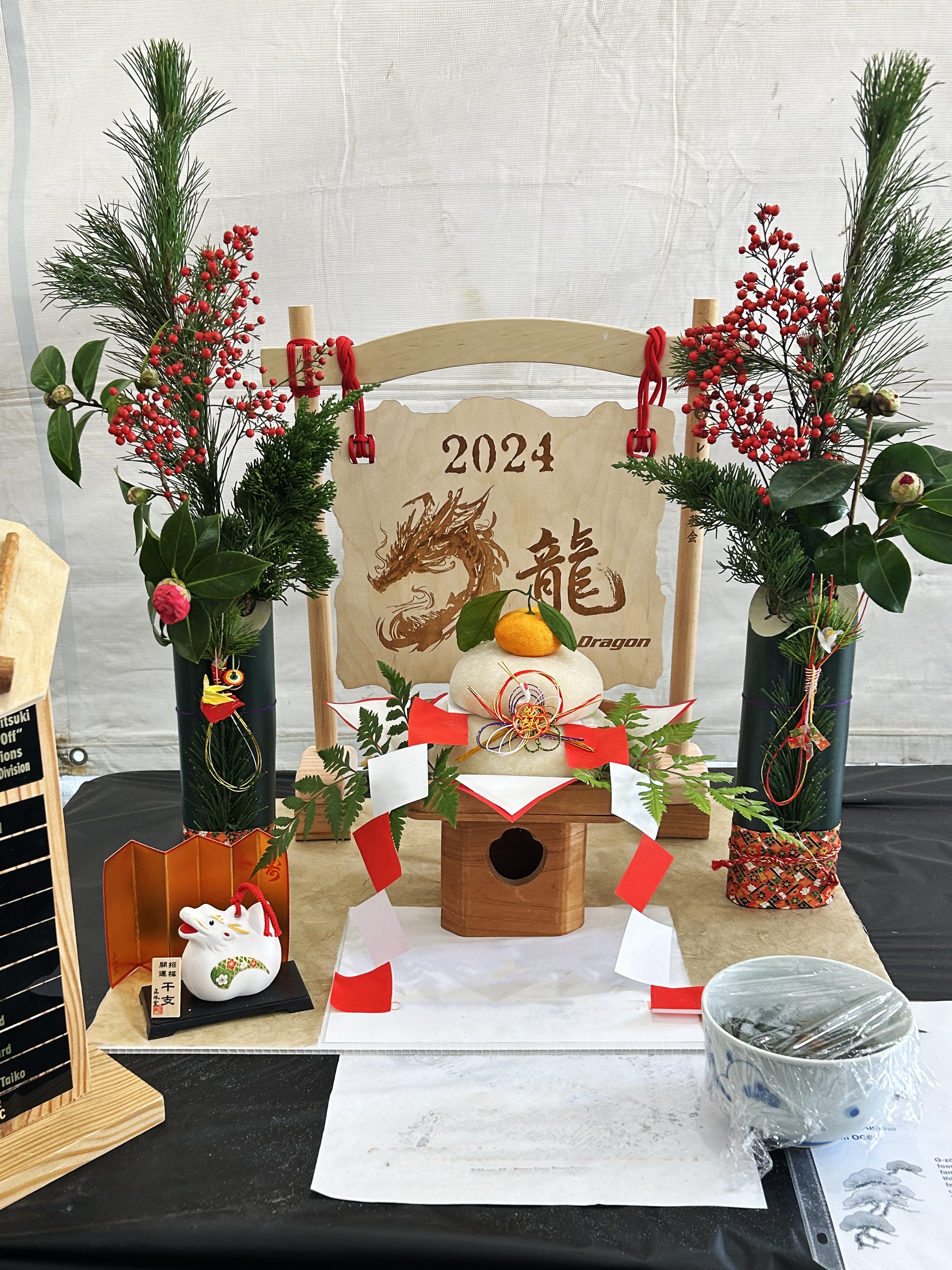 This image features a Japanese New Year decoration display with a small wooden shrine, a kagami mochi (stacked rice cakes with a citrus fruit on top), and traditional floral arrangements with red berries and green foliage. There is also a white ceramic fox figurine and a small round glass bowl on the table in front.