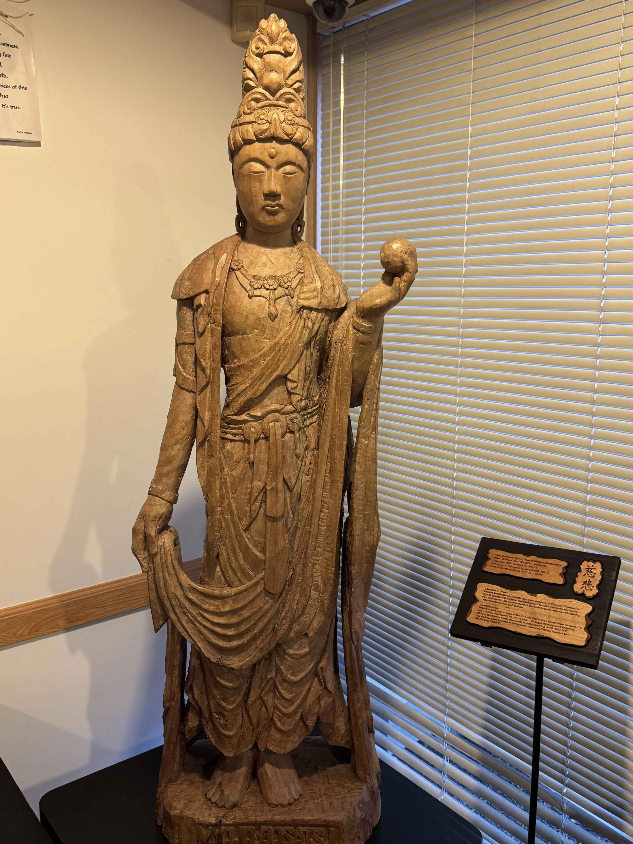 Wooden carved statue of a female figure with a headdress and jewelry, holding an orb in her right hand, displayed indoors near window blinds and informational plaque.
