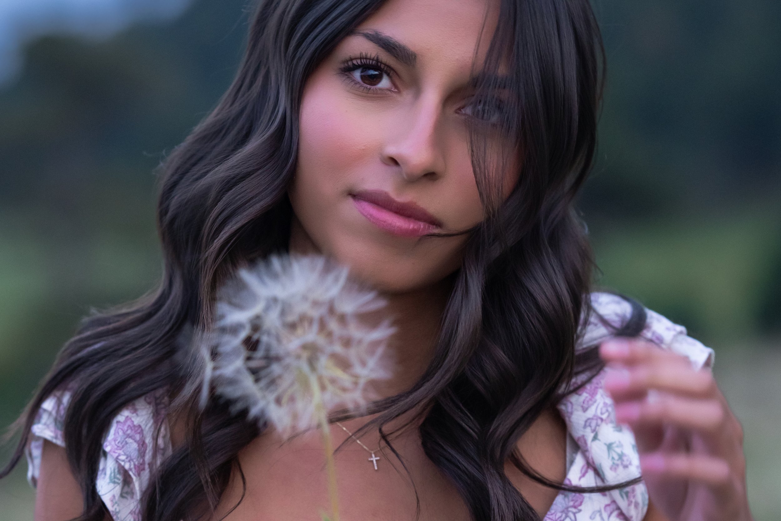 Close-up of a young woman with dark wavy hair and brown eyes, holding a dandelion seed head, outdoors with a blurred background.