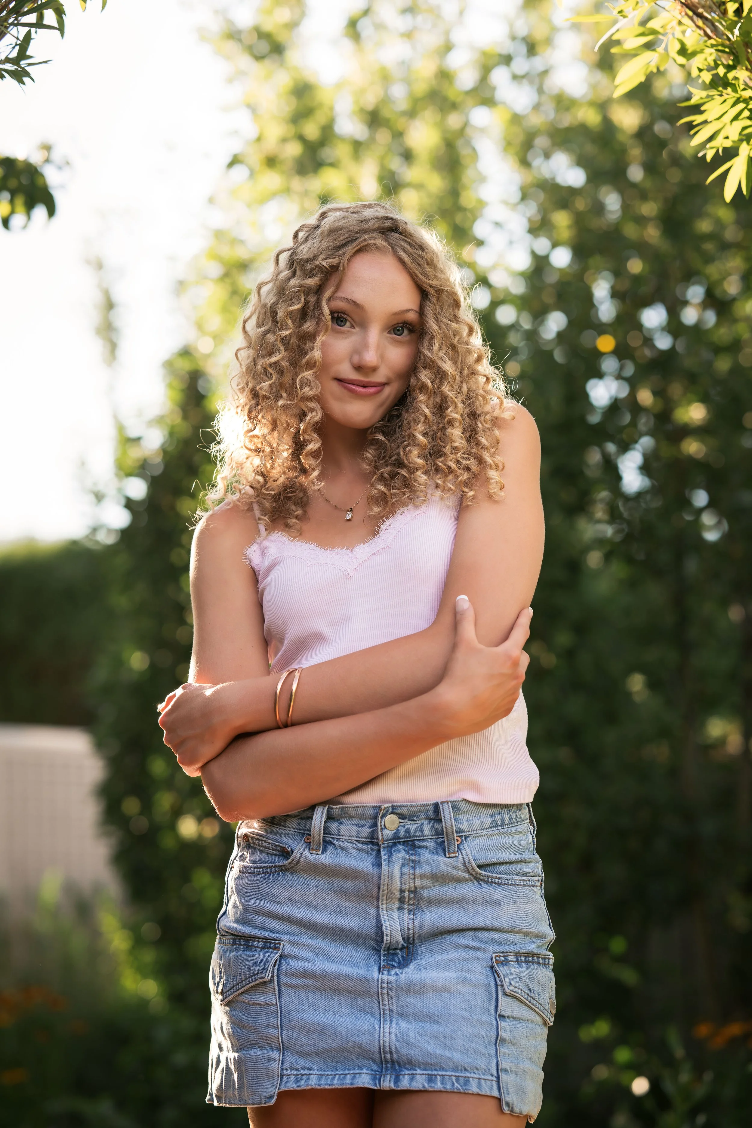 Young woman with curly blonde hair wearing a light cami top and jean skirt poses with her arms crossed.