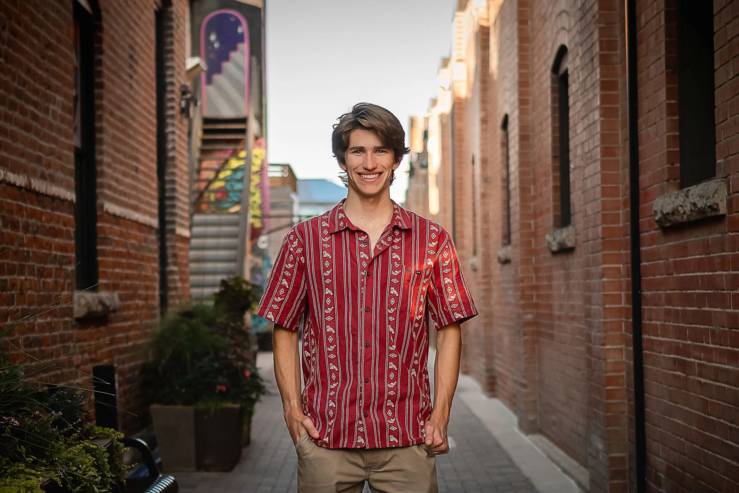 Young man walking down a brick alleyway, smiling, wearing a red and white patterned short-sleeve shirt and beige shorts, with buildings and greenery on either side.