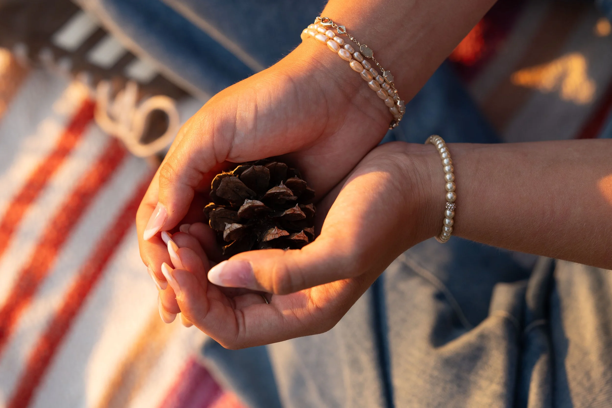 A girl with a French manicure holds a pinecone in her hand and has a pearl bracelet on one arm and stacked bracelets on the other hand.