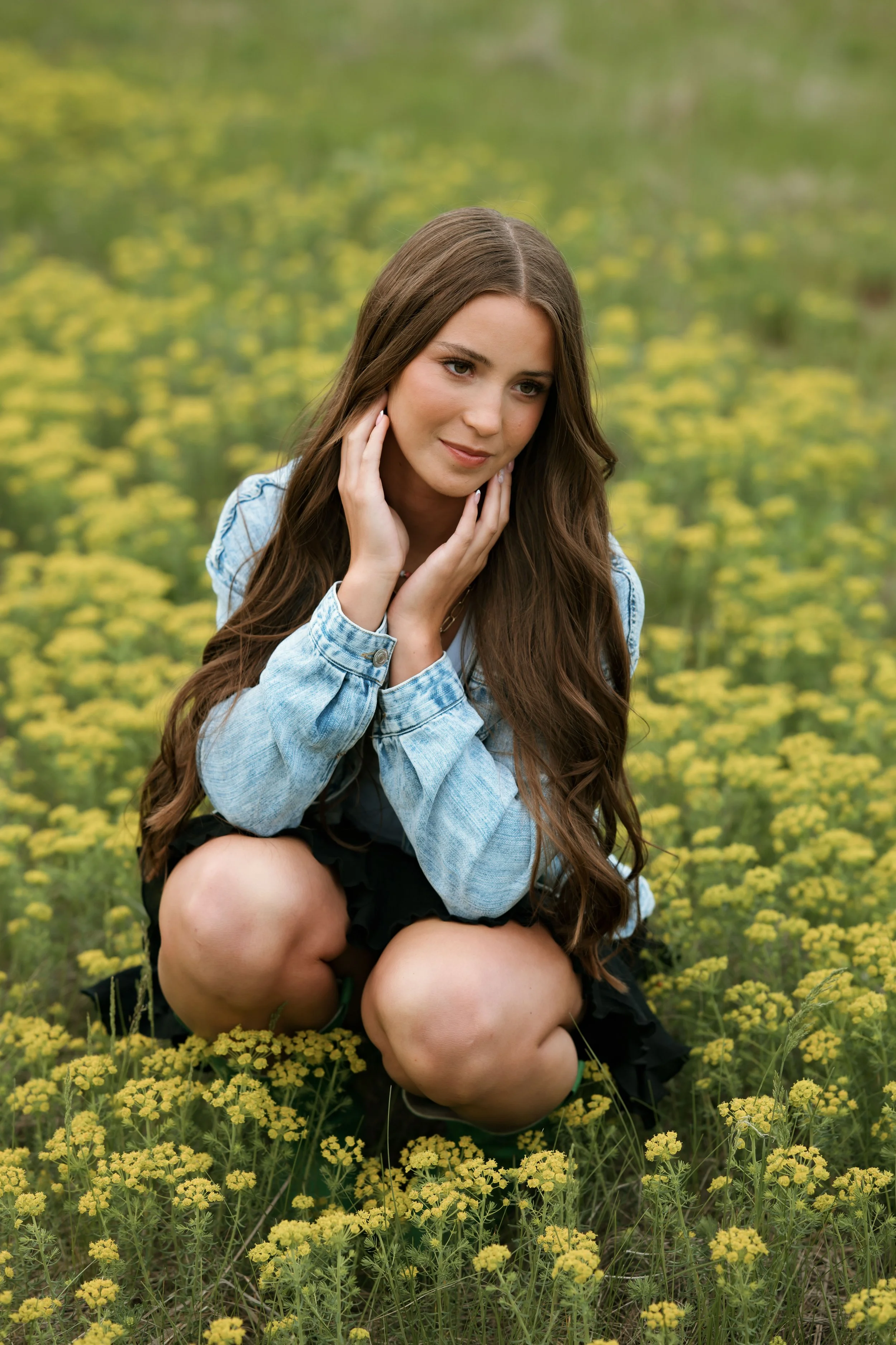 A young woman with long brown hair, wearing a denim jacket and black shorts, is kneeling in a field of yellow flowers, gently touching her face with her hands and looking thoughtful.
