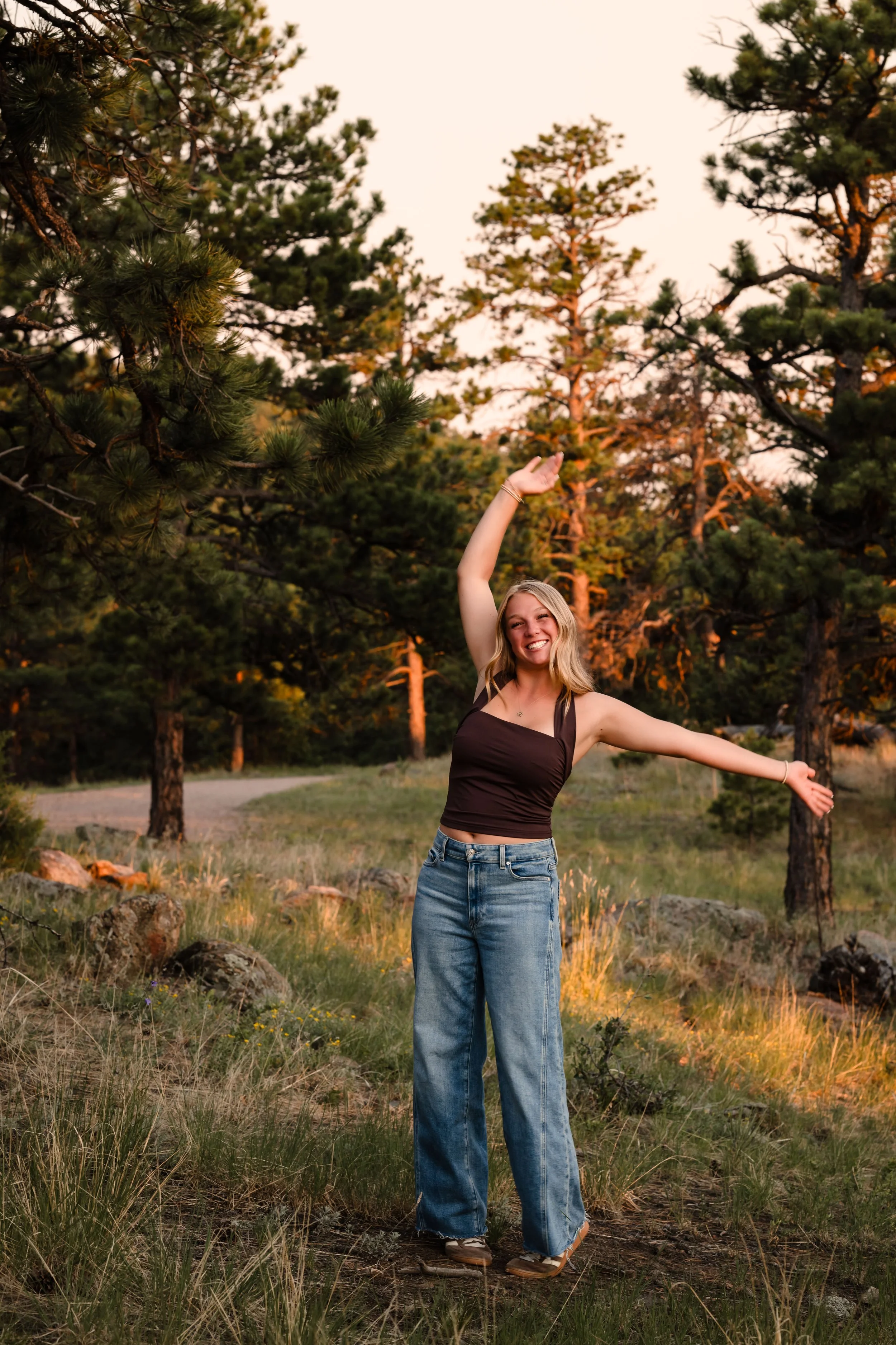 Blonde girl with a brown tank top and jeans poses with one arm up in the air and the other straight out. She has a big smile.