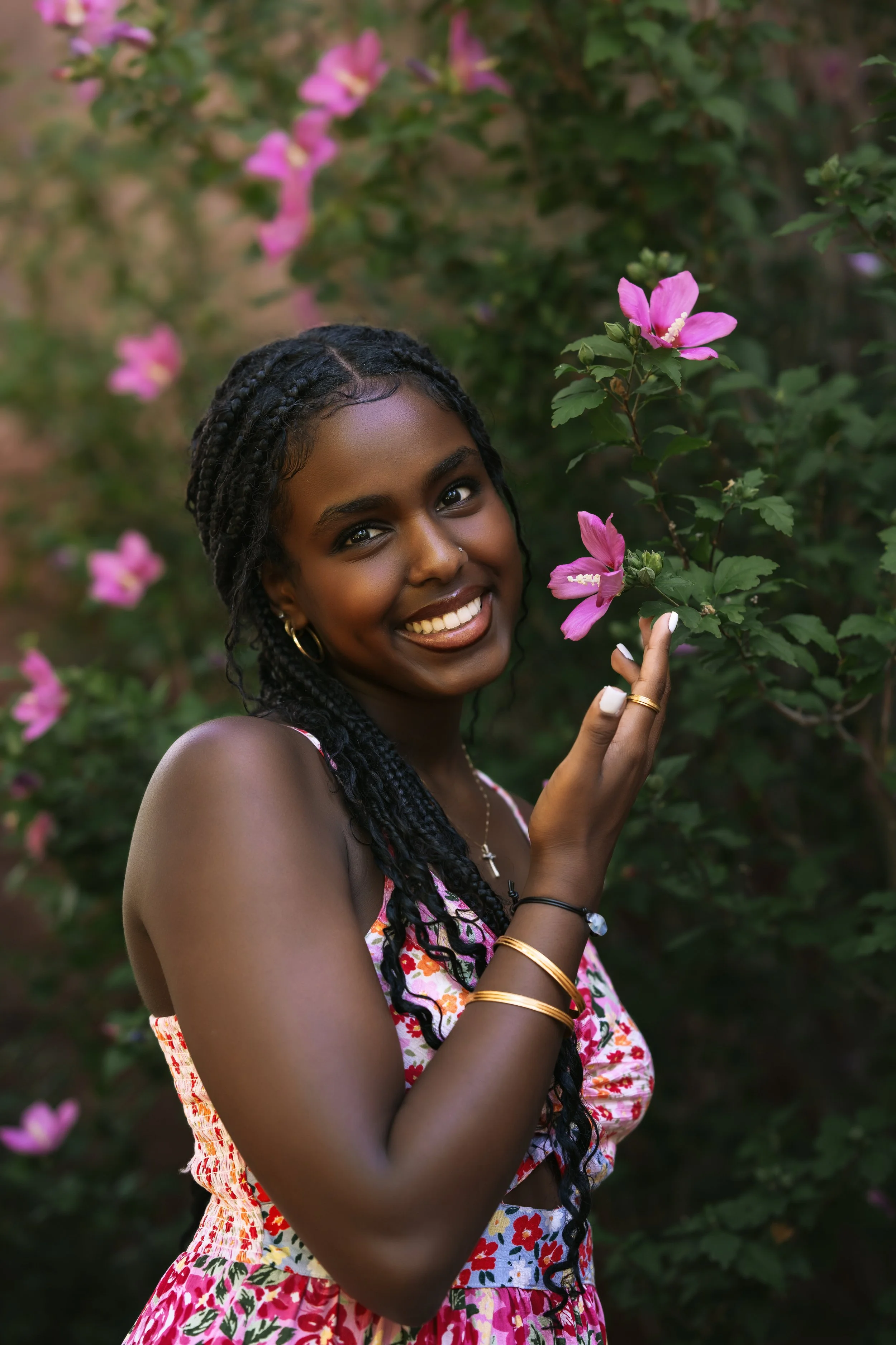 African American teen wearing a pink floral dress smells a pink floral bush at University of Denver for her senior pictures. 