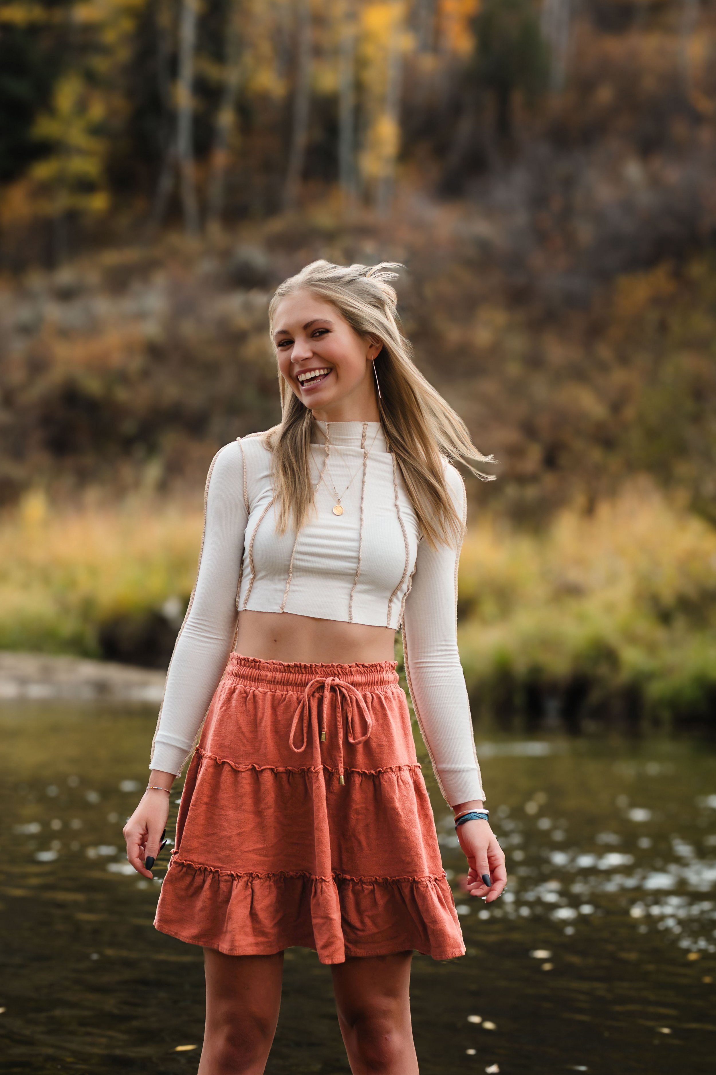 A young woman with long blonde hair, smiling, wearing a white long-sleeve crop top and a rust-colored, tiered skirt, standing near a body of water with trees and fall foliage in the background.
