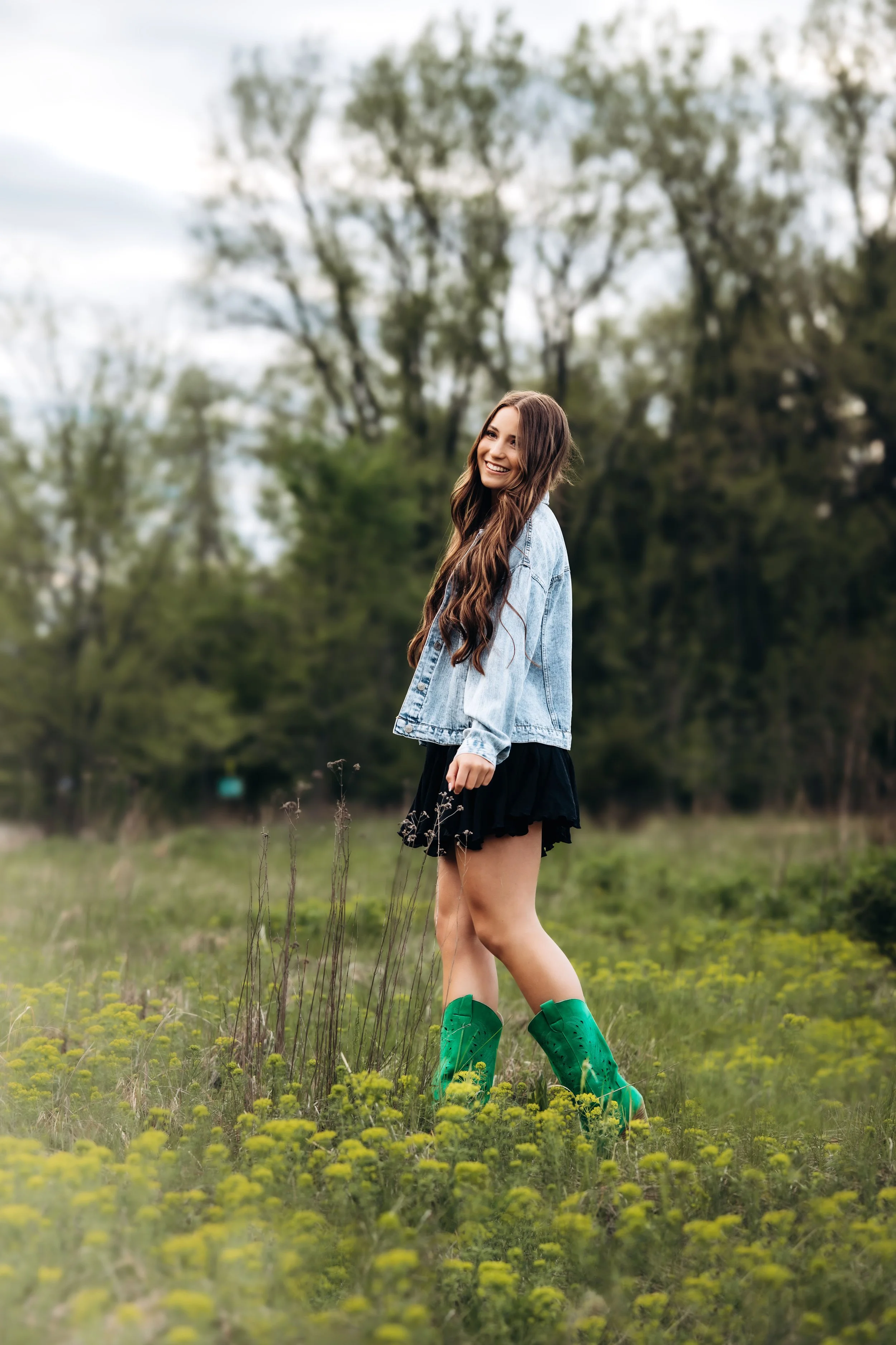 A young woman smiling and standing in a grassy field with yellow flowers, wearing a denim jacket, black skirt, and green rain boots, with trees and a cloudy sky in the background.