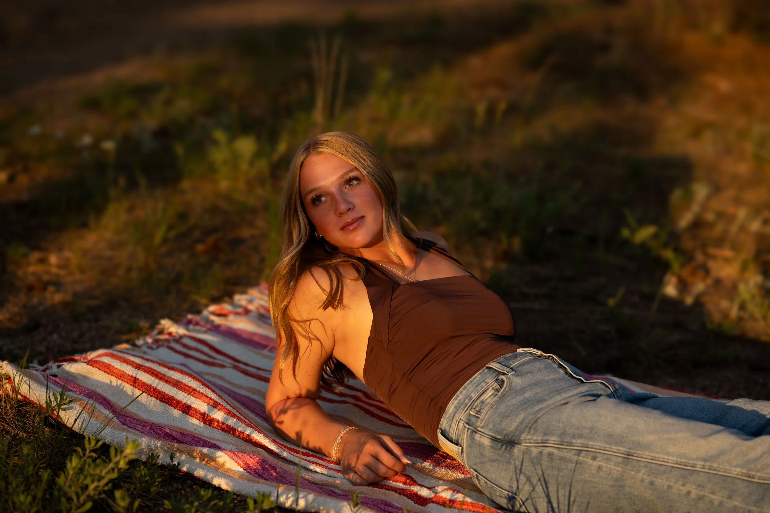 Blonde girl lays back on a striped picnic blanket near some pine trees. She looks off into the distance and poses for her senior pictures.