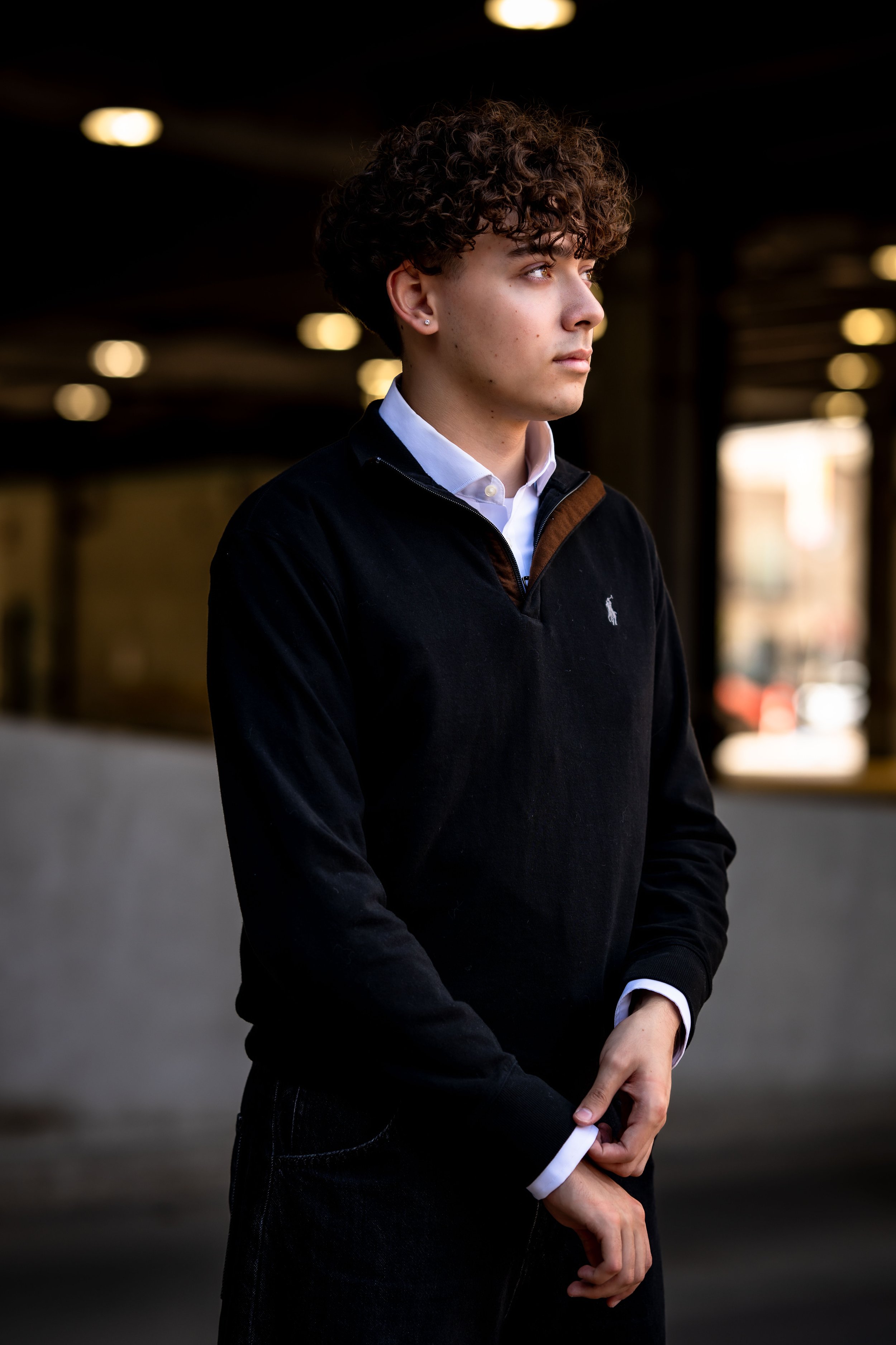 A young man with curly hair and earrings wearing a black sweater over a white collared shirt, standing outdoors in a dimly lit urban area at night, looking to the right.