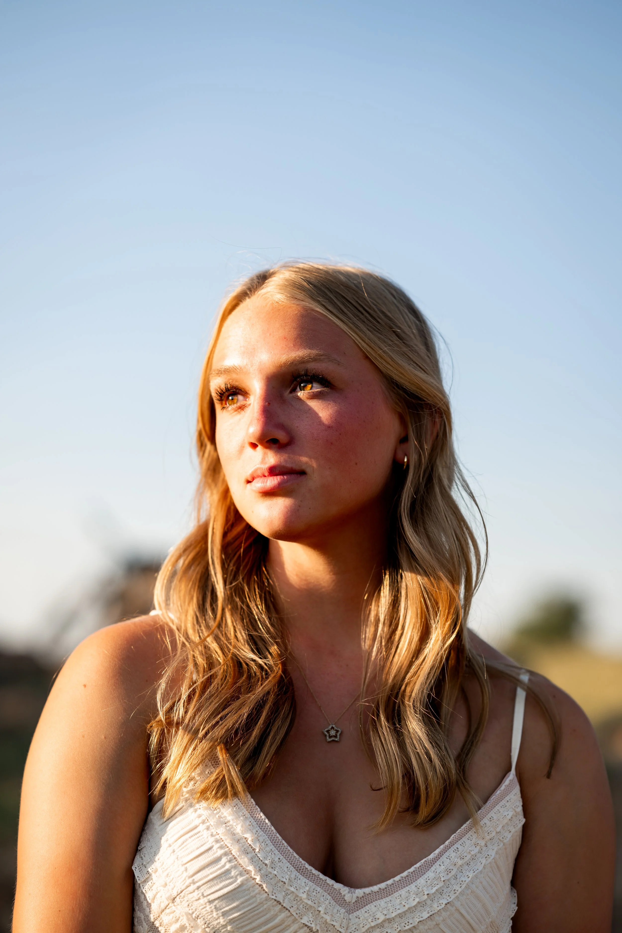 Blonde girl with brown eyes wears a white v-neck dress and looks toward the sun.