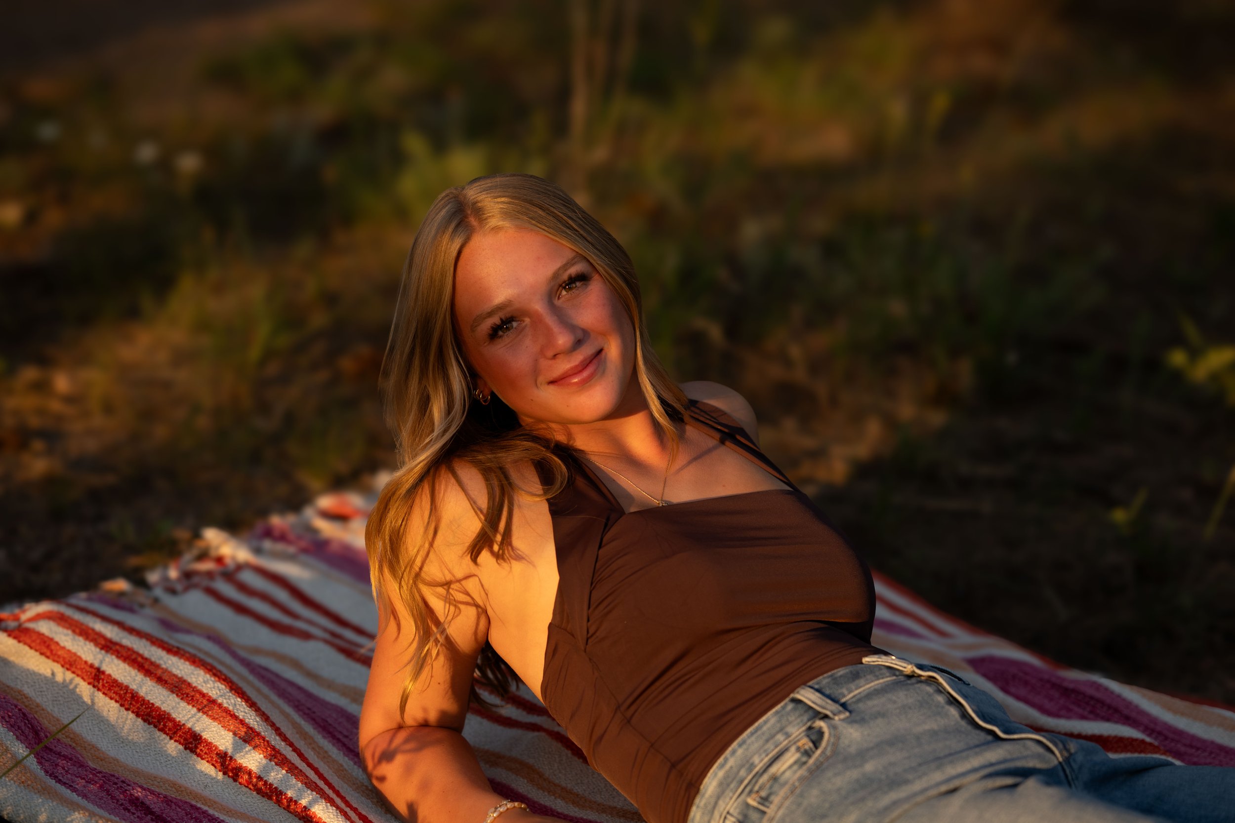 High School senior lays on a striped picnic blanket in the pine trees with some soft light on her face.