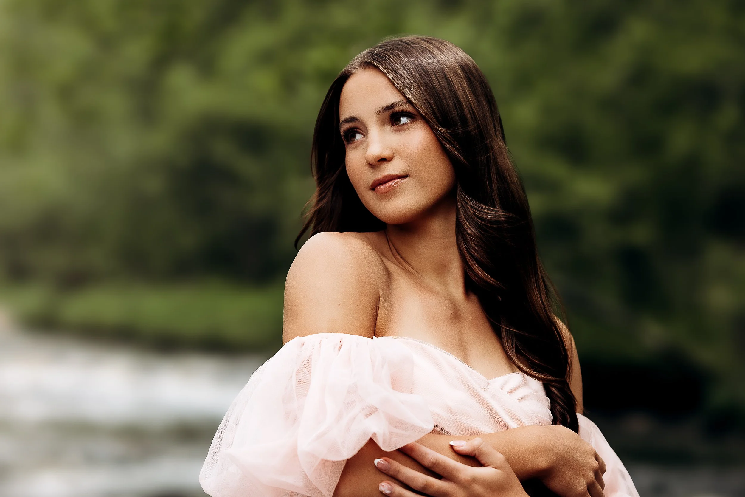 A woman with long dark hair stands outdoors with her arms crossed, wearing a light-colored off-the-shoulder dress, and looking to the side with a thoughtful expression, blurred green foliage in the background.