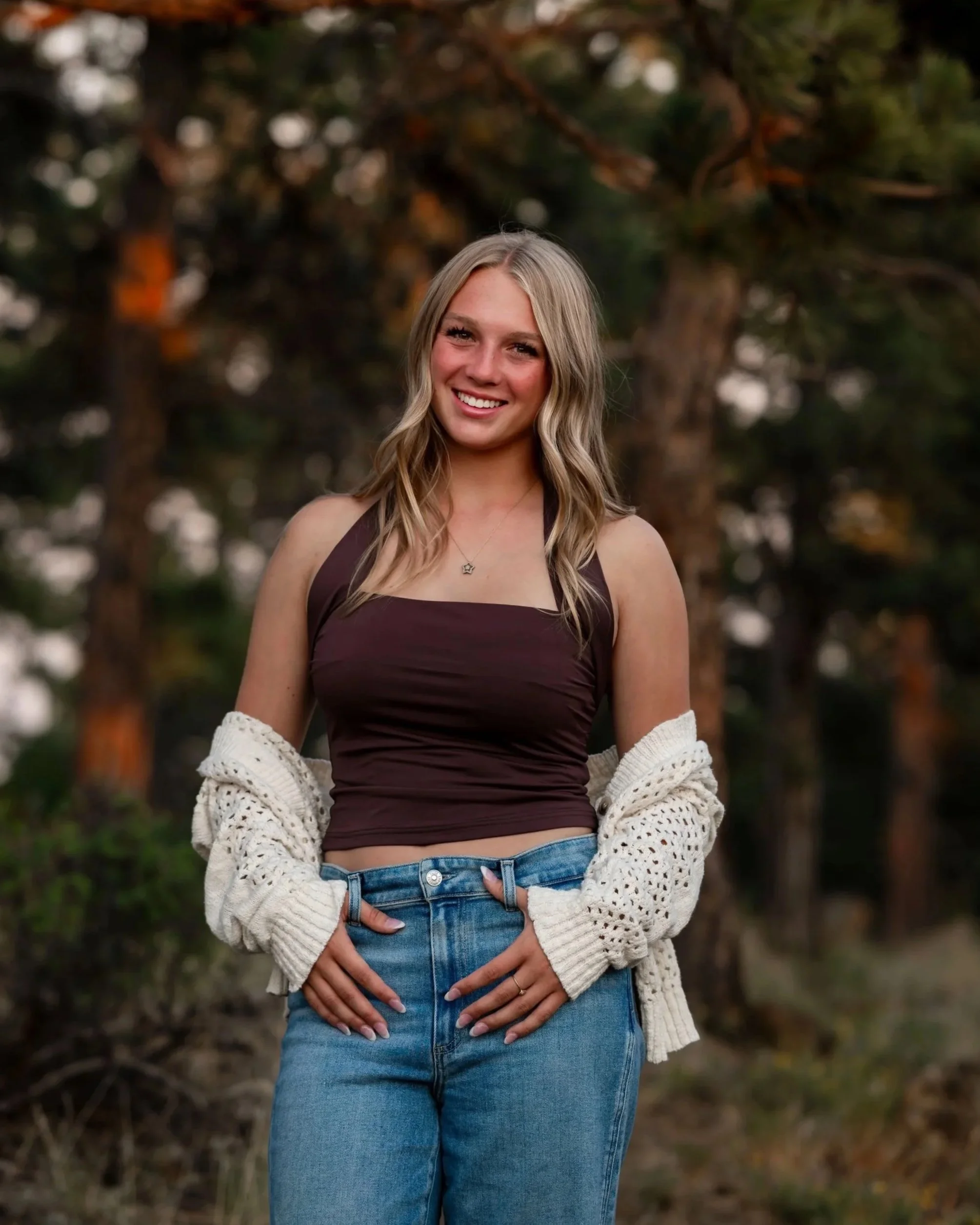 A young woman smiling outdoors with trees in the background, wearing a dark brown tank top, a white knitted cardigan hanging off her shoulders, and blue jeans. She has long, wavy blonde hair and a necklace with a star-shaped pendant.